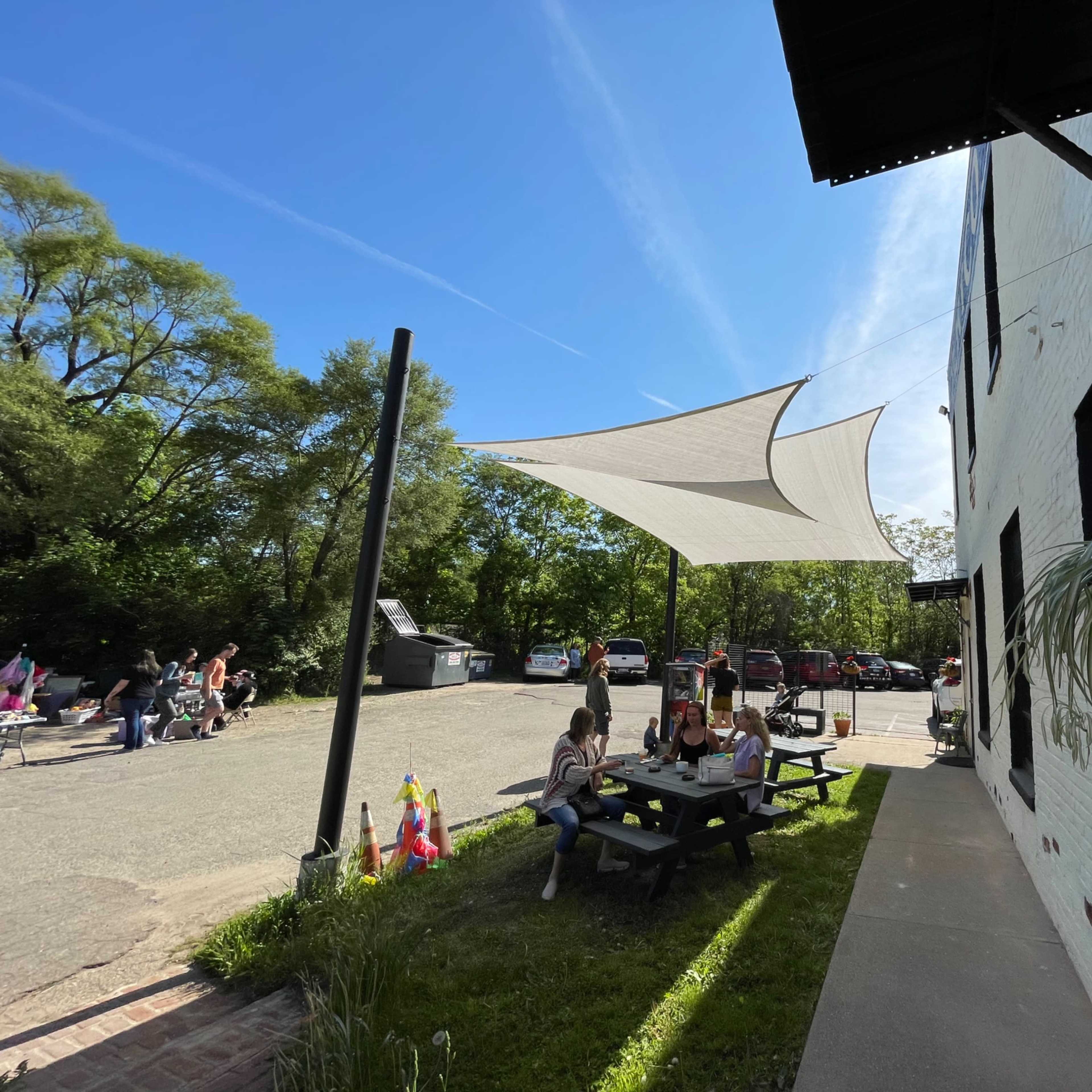 A shaded outdoor seating area with a picnic table is filled with people beside a parking lot surrounded by trees.