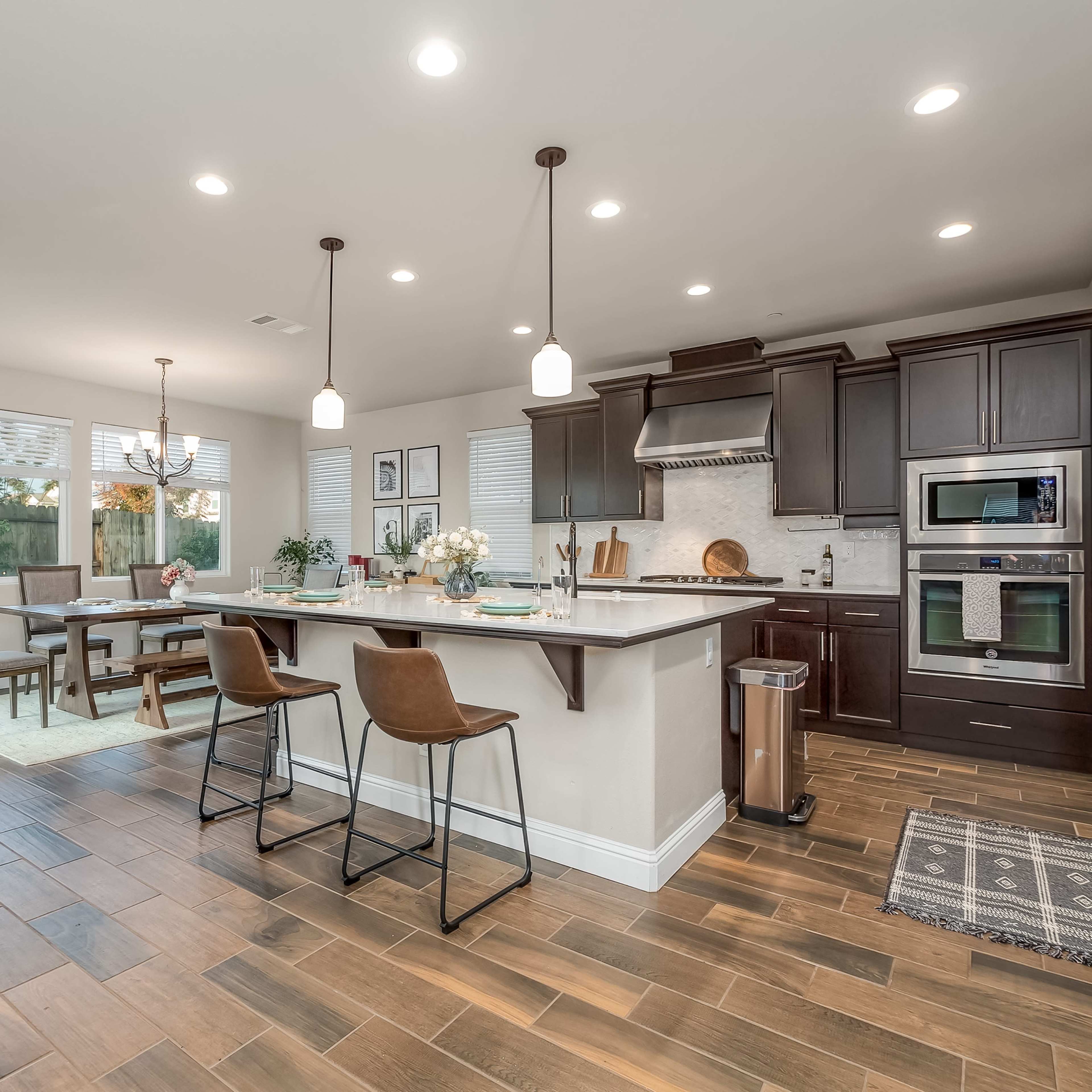 A modern kitchen with dark wood cabinets, a central island with bar stools, and a dining area featuring a table and chairs, all illuminated by overhead lights.