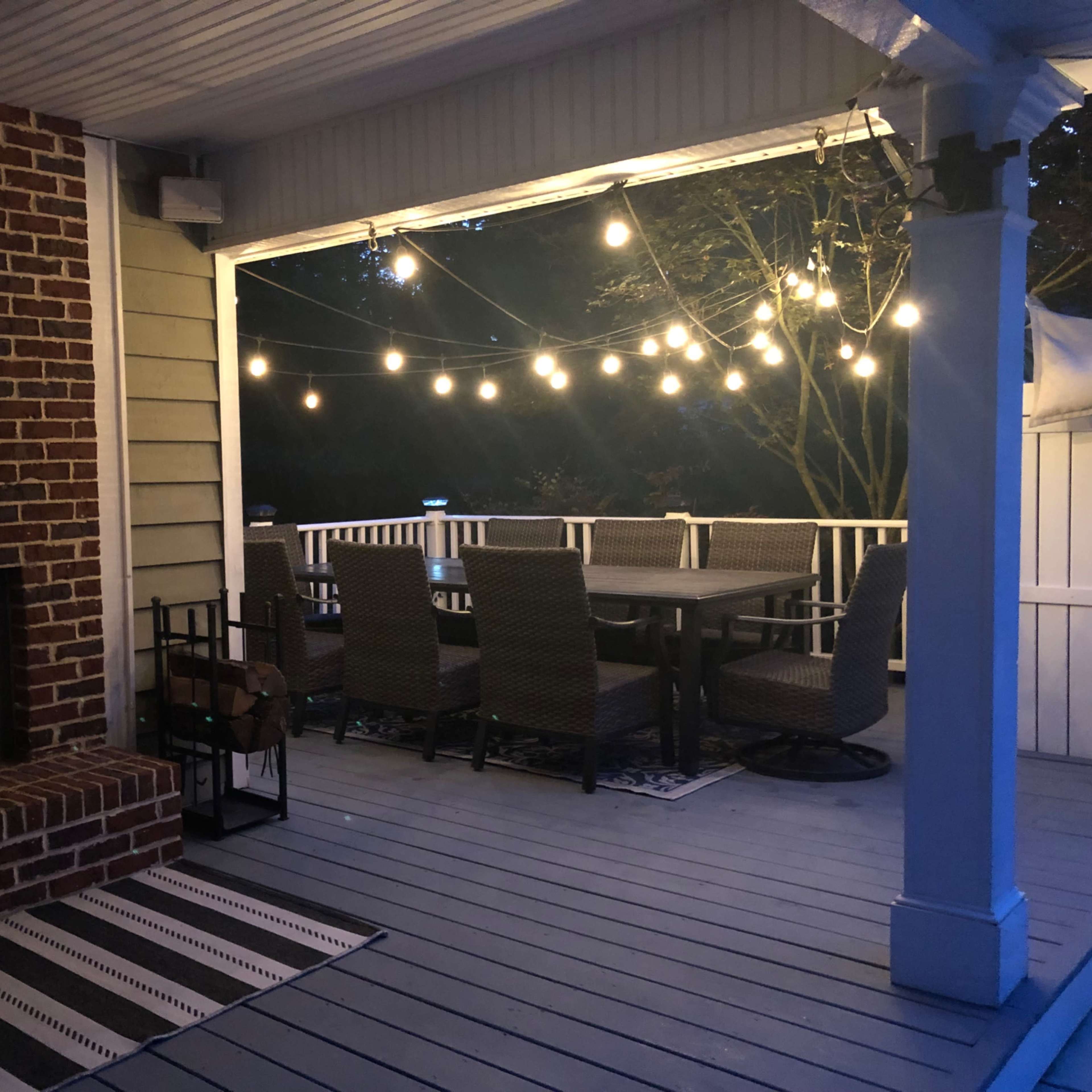 A covered outdoor patio area featuring a brick fireplace with candles, a large dining table surrounded by wicker chairs, and string lights overhead.
