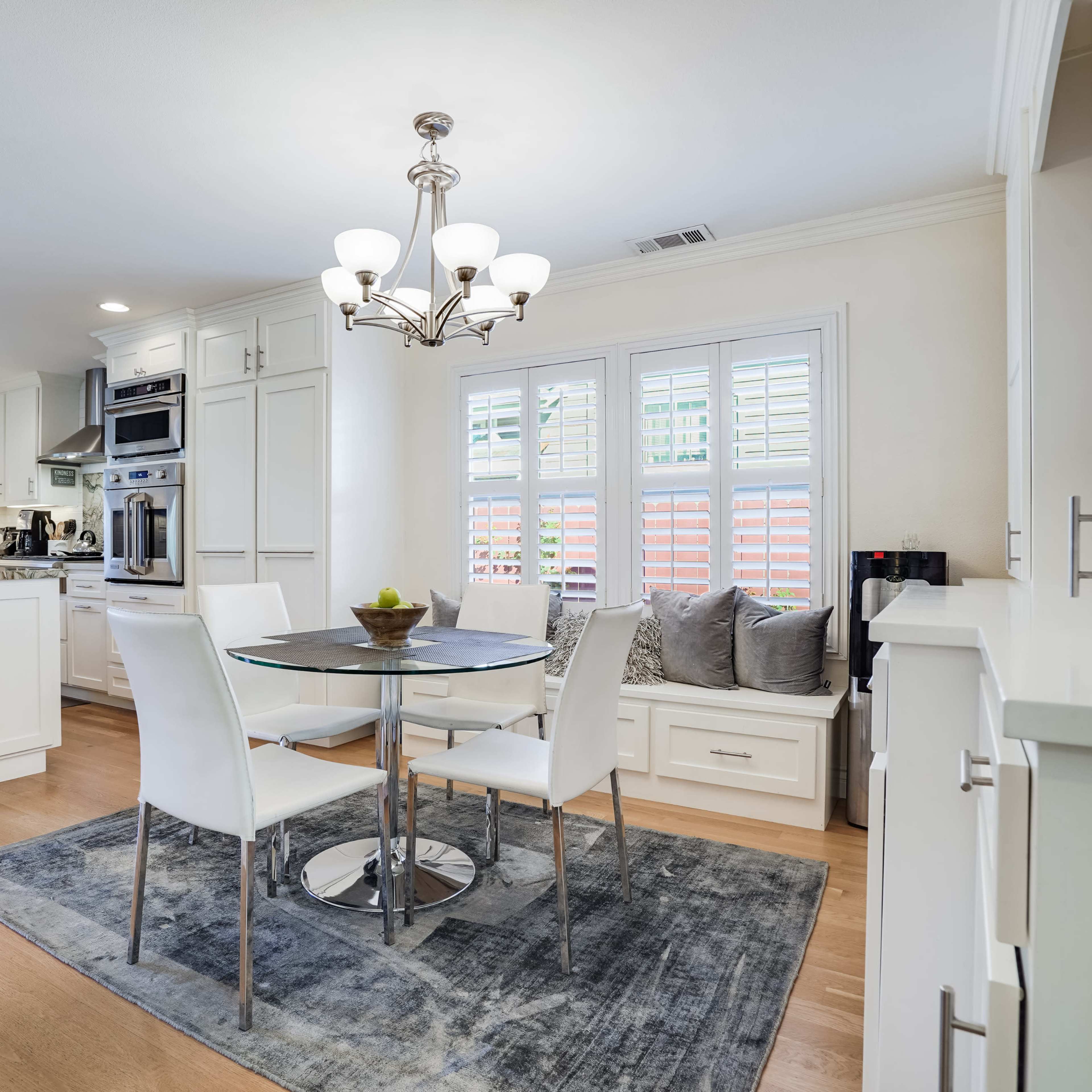 A modern kitchen and dining area features a round glass table surrounded by white chairs, with a chandelier above and a window displaying plants outside.