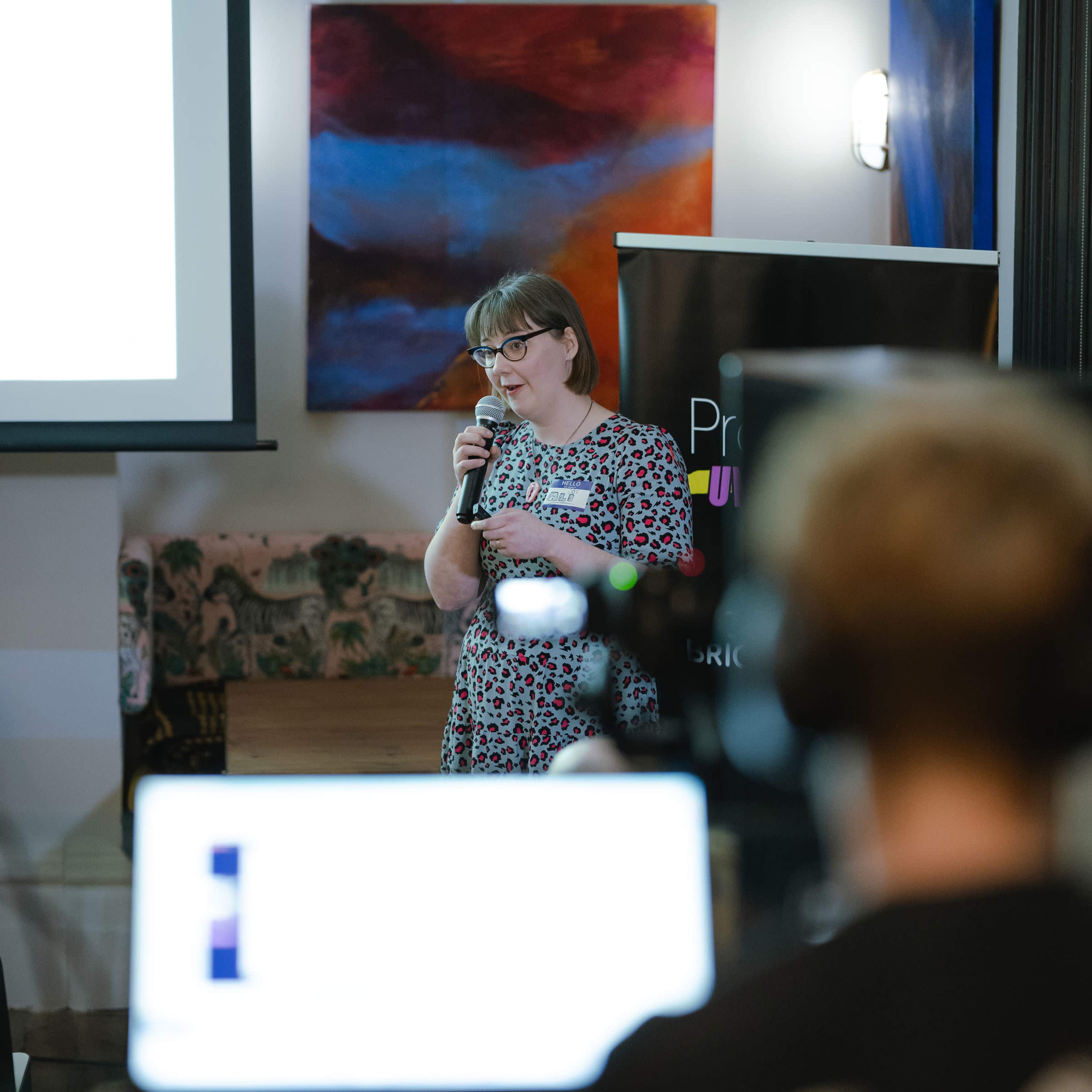 A woman speaks into a microphone in front of a presentation screen, while a blurred laptop and audience are visible in the foreground.
