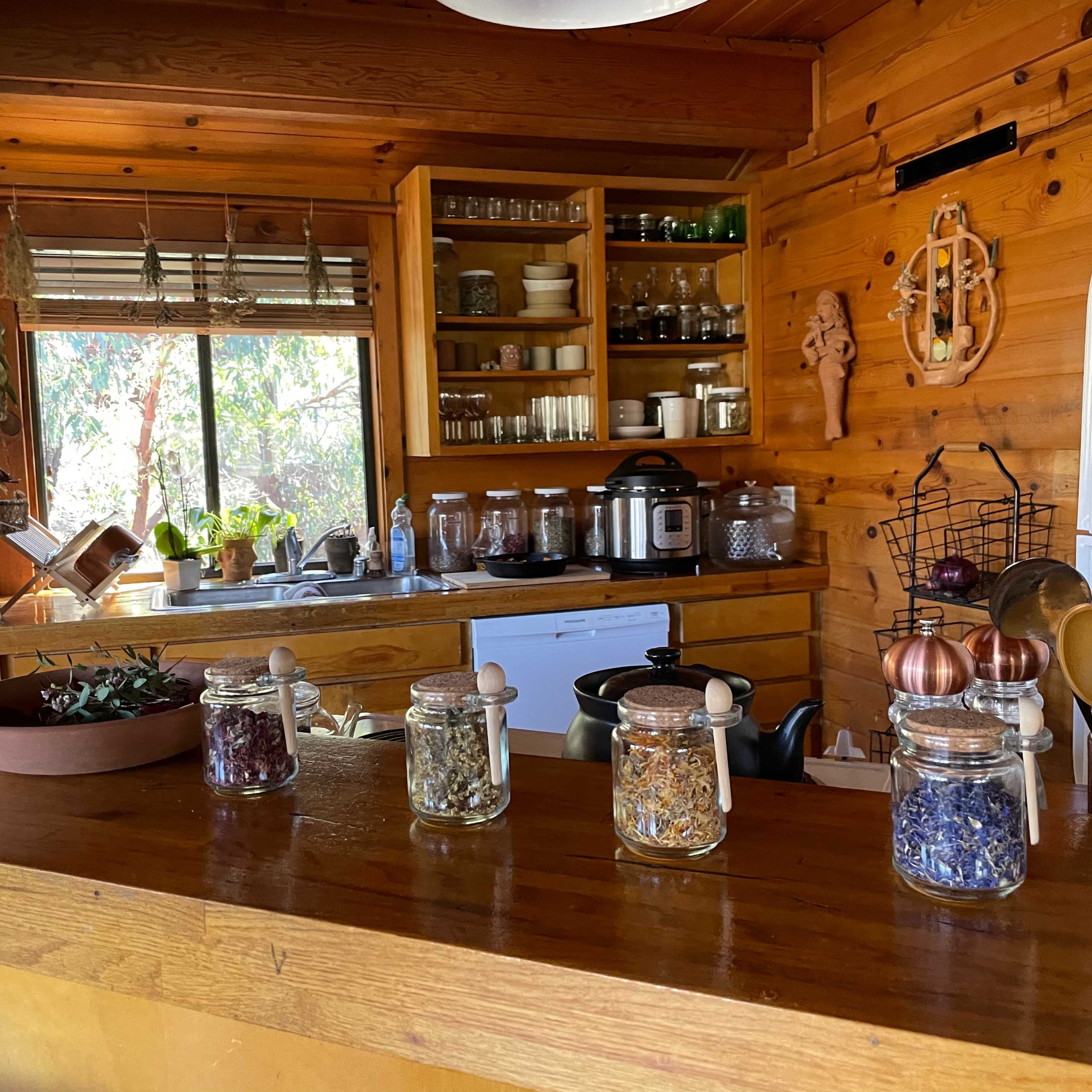 A wooden kitchen features jars of herbs lined up on a counter, with shelves of glassware and cooking utensils visible in the background.