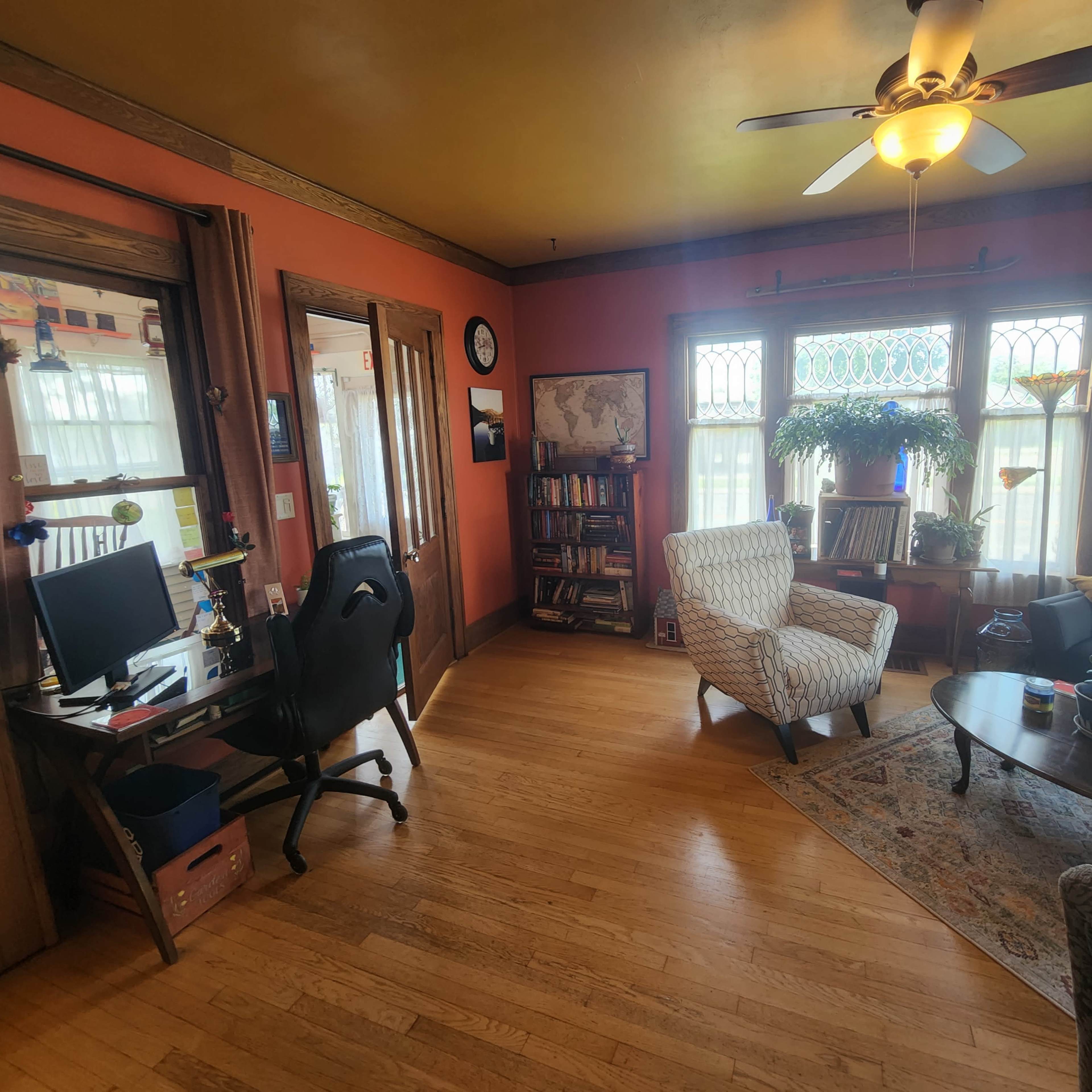 A cozy living room with a desk and computer, two armchairs, a table, and large windows allowing natural light to enter.