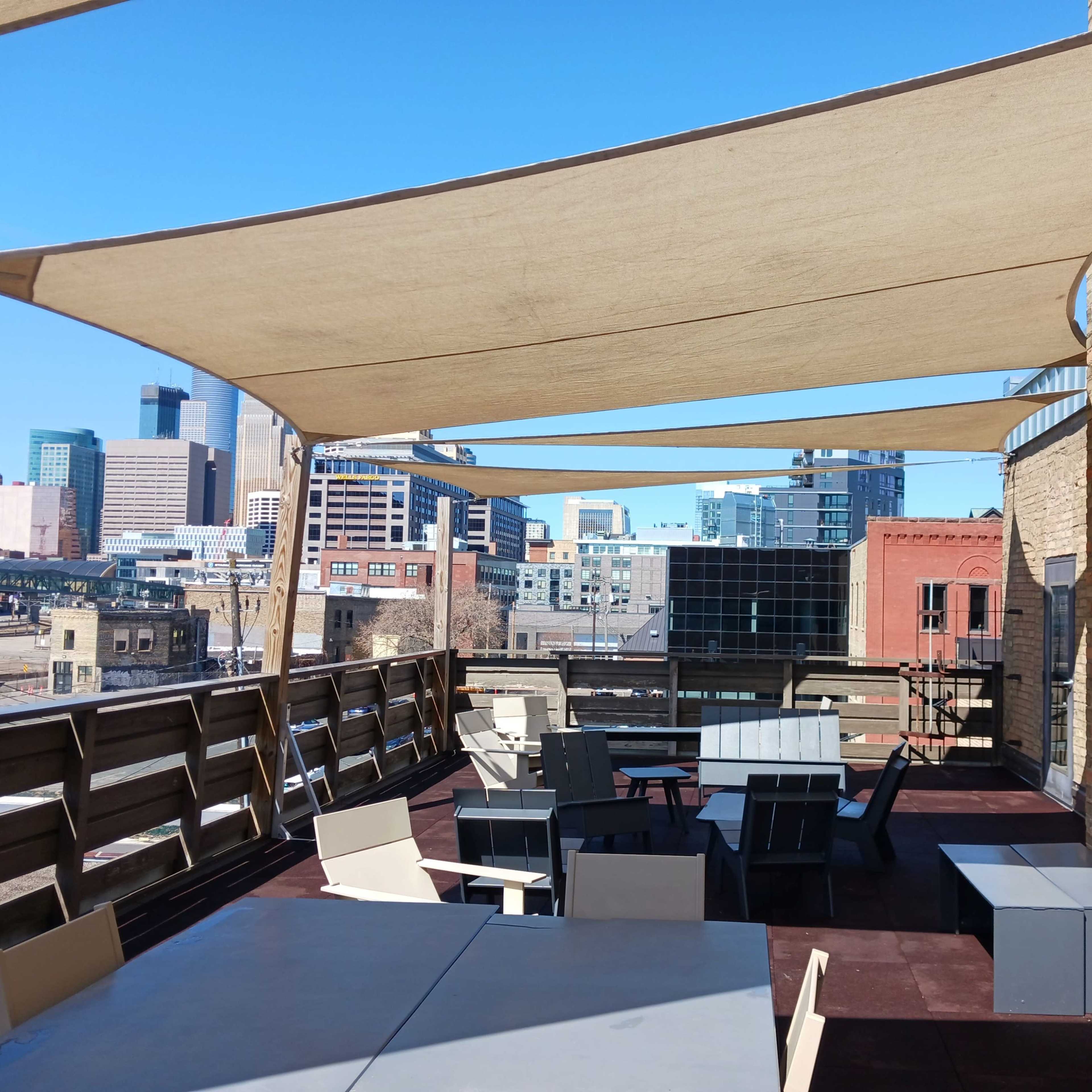 A rooftop terrace with several tables and chairs shaded by large fabric canopies, overlooking a city skyline.