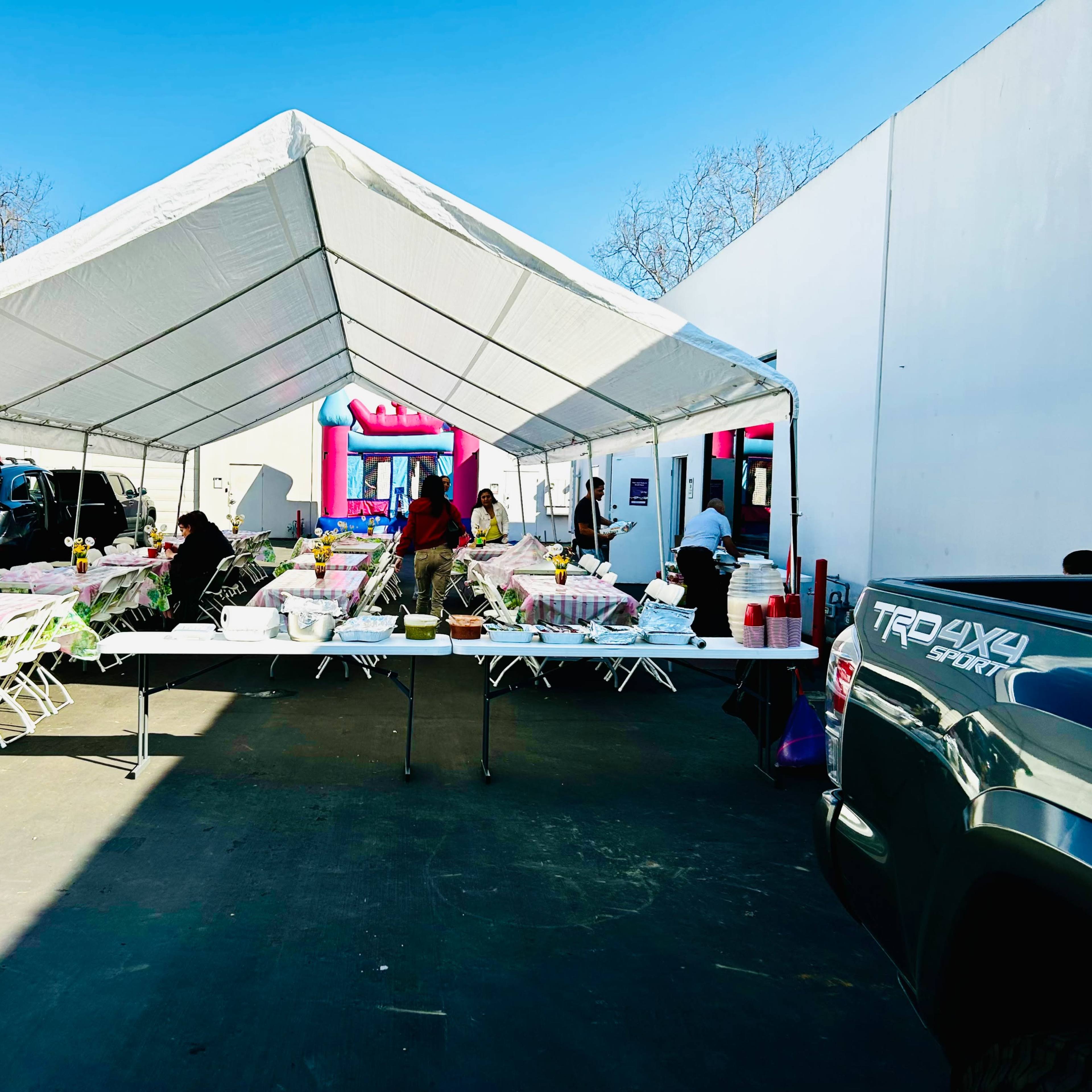 A large white tent is set up in a parking lot, filled with tables covered in checkered cloths and catered food, while people gather around, and a bounce house can be seen in the background.
