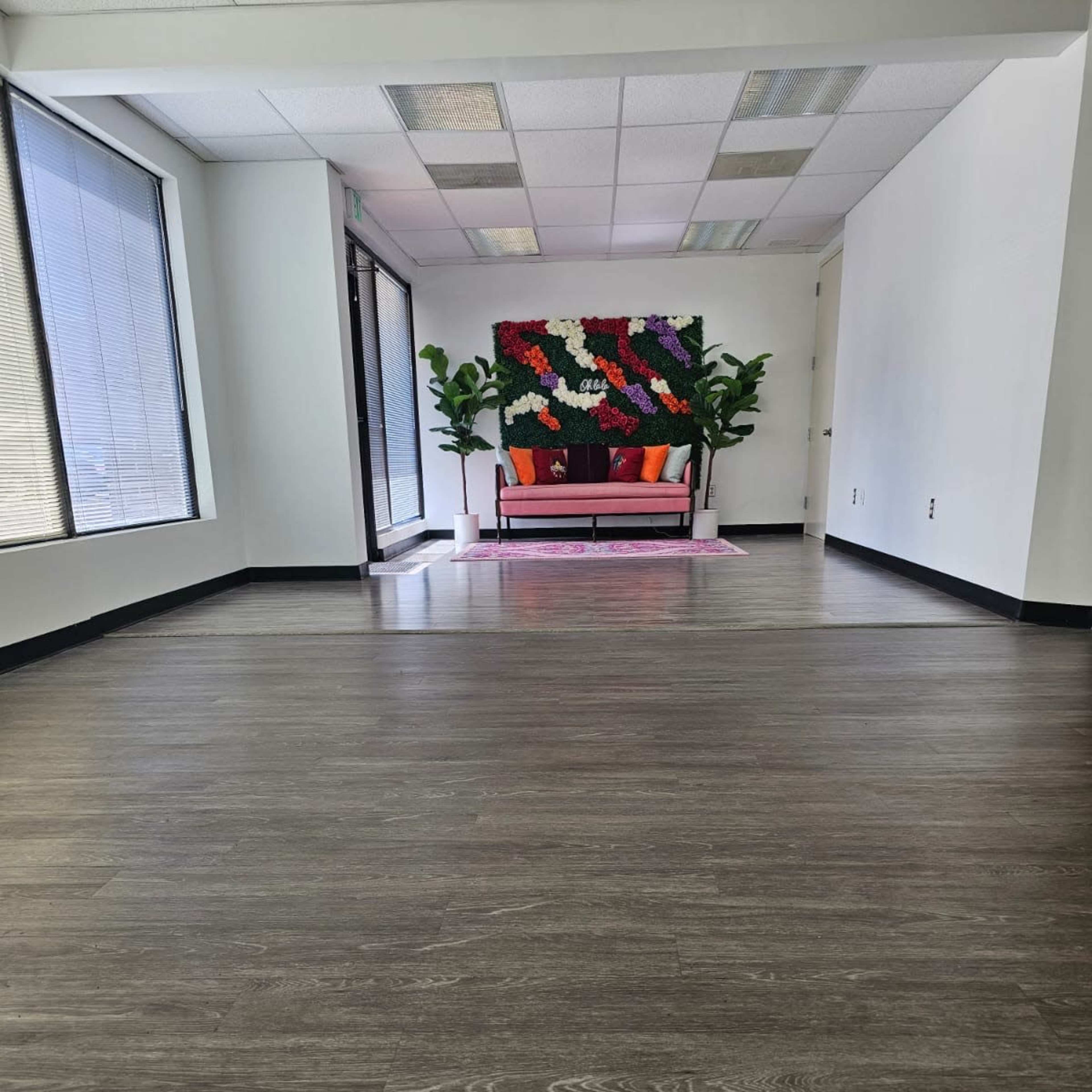 An empty room with dark wooden flooring, large windows, and a colorful floral backdrop behind a pink sofa.