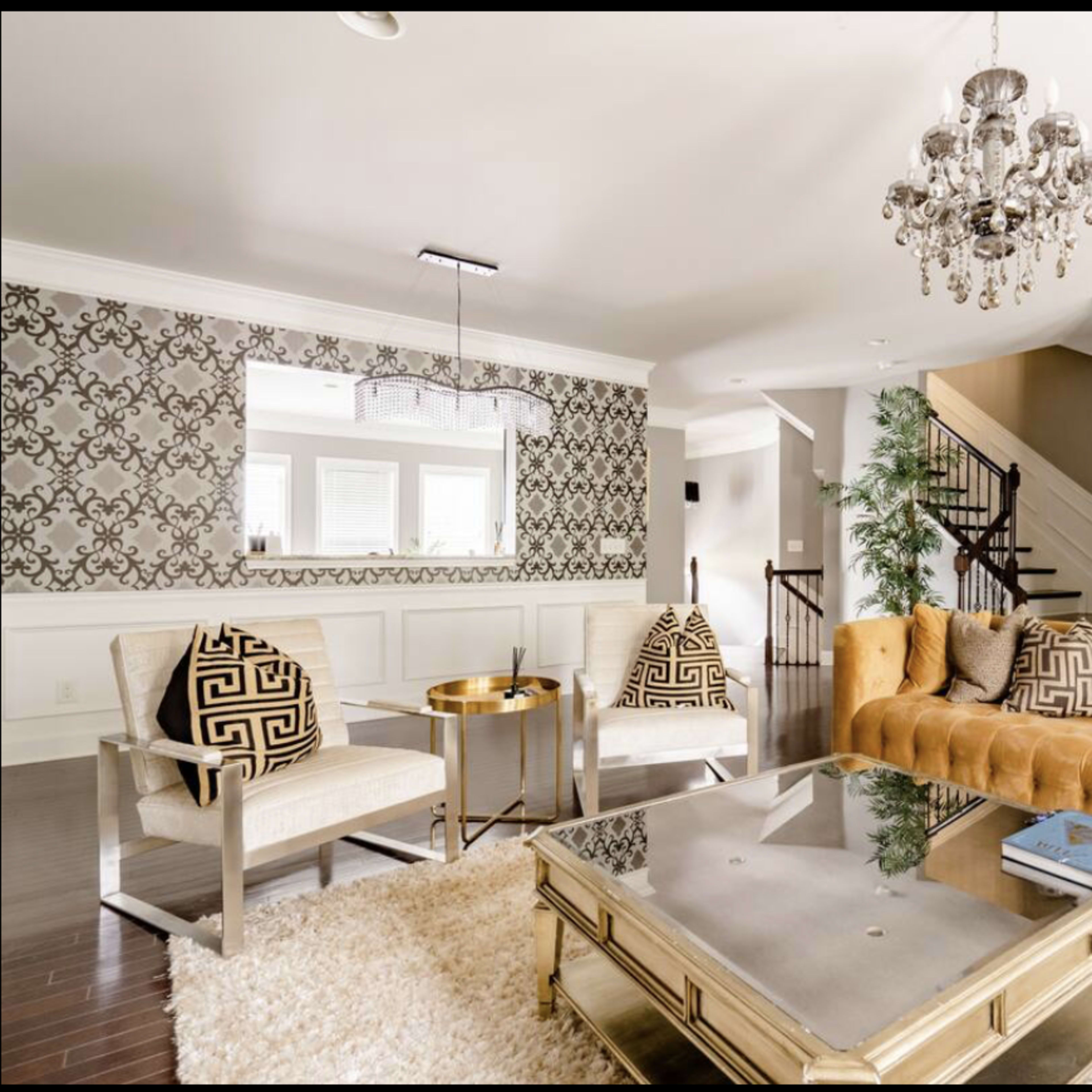 A well-furnished living room features a tan tufted sofa, two patterned armchairs, a glass coffee table, and a chandelier, with decorative wallpaper and hardwood flooring.