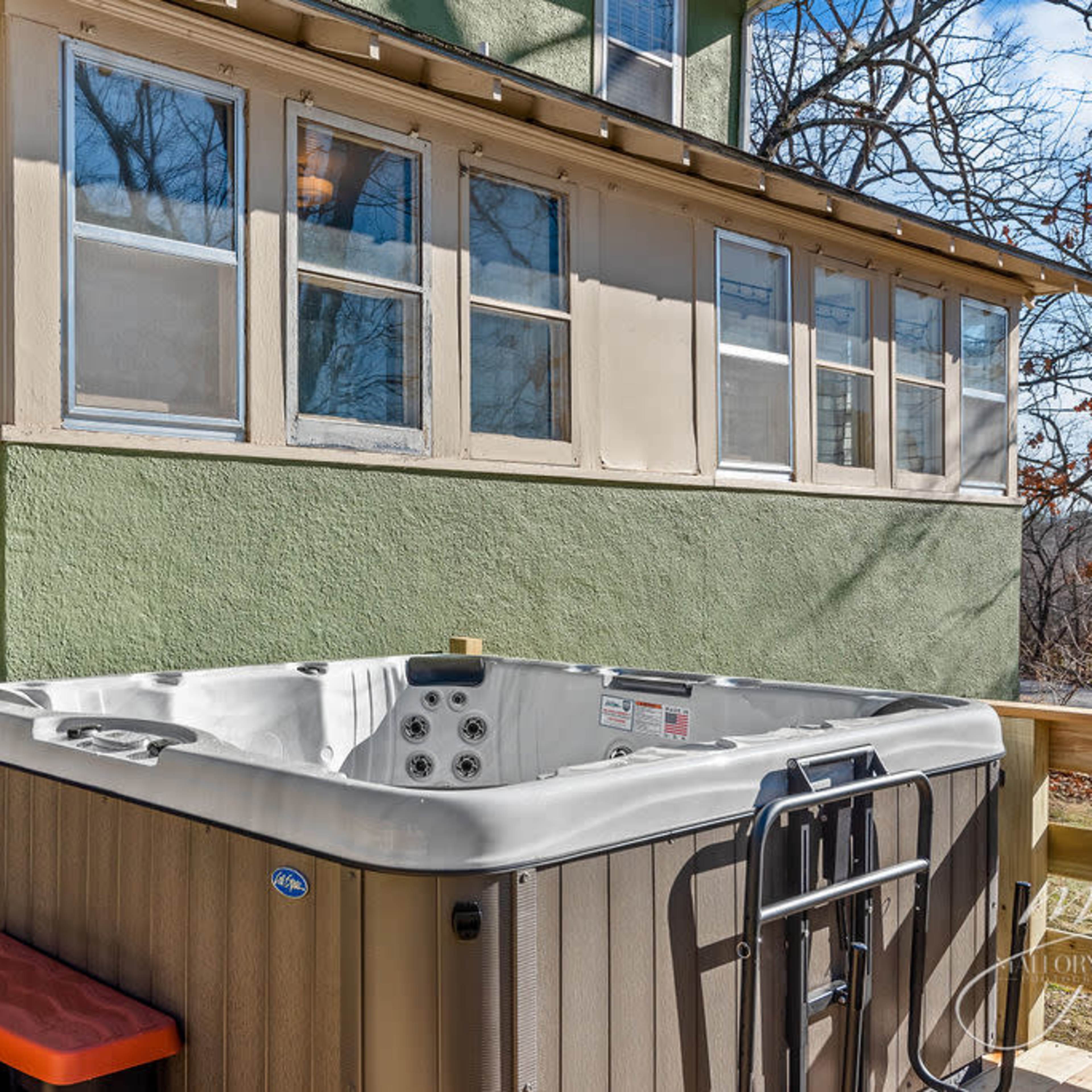 A hot tub positioned on a wooden deck beside a green house with multiple windows.