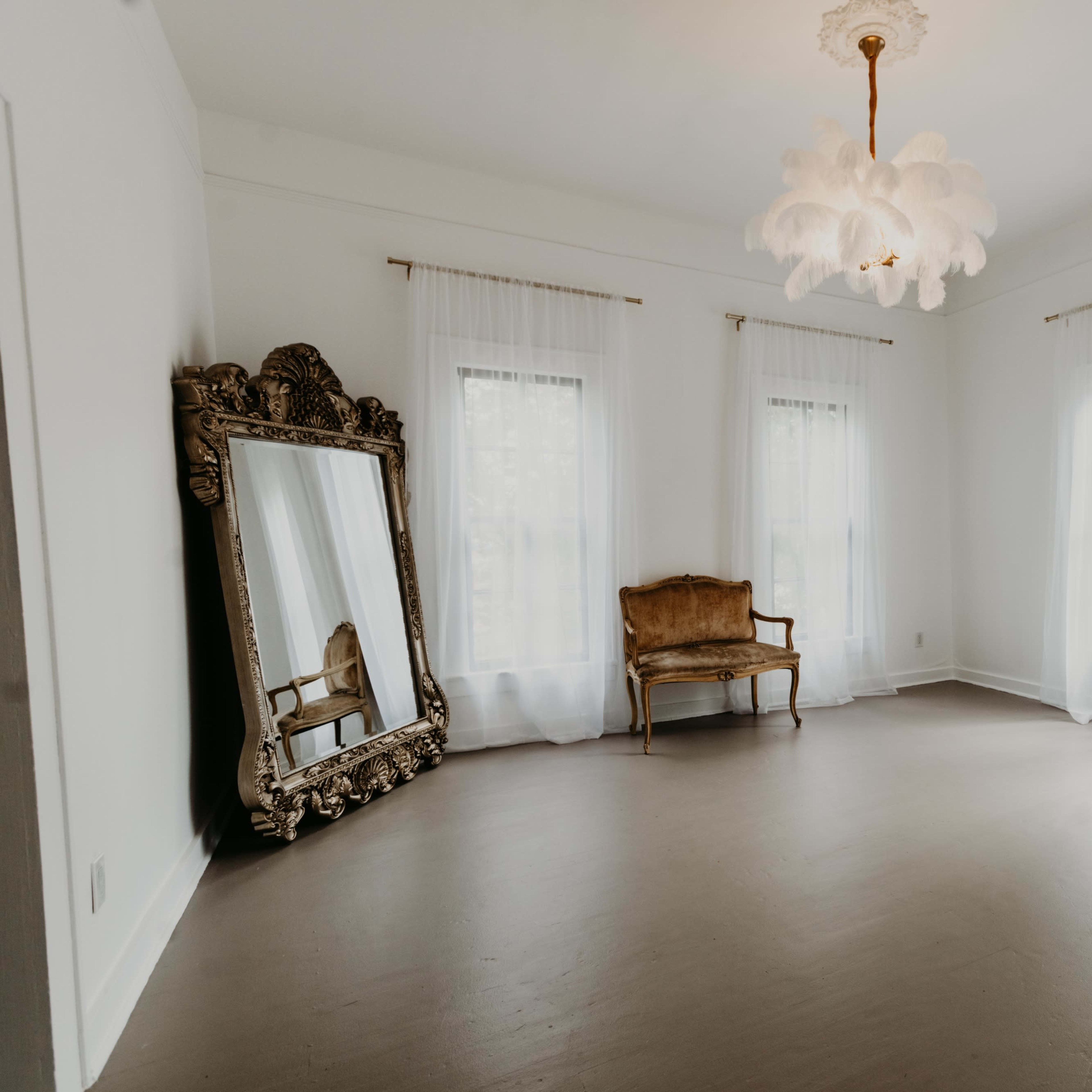 An empty room featuring two large windows with sheer curtains, a decorative mirror, a vintage armchair, and a chandelier.
