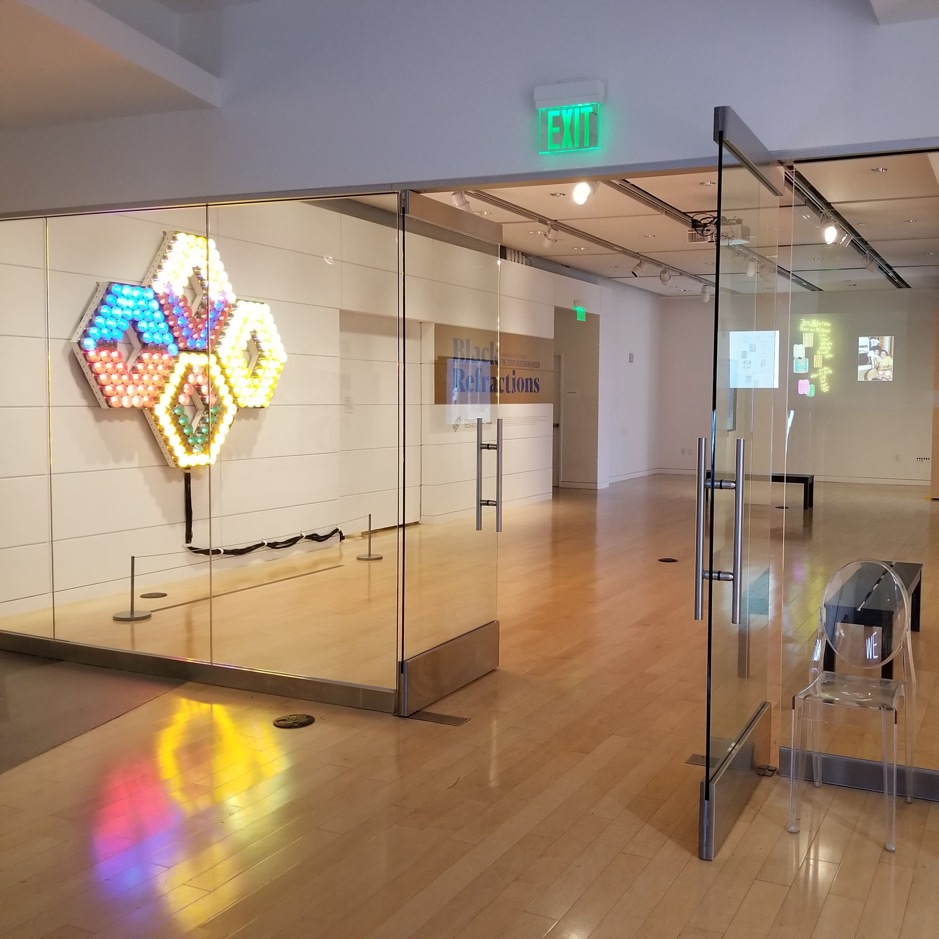A modern art gallery with a colorful hexagonal light installation on one wall and glass doors leading to the exhibition space.