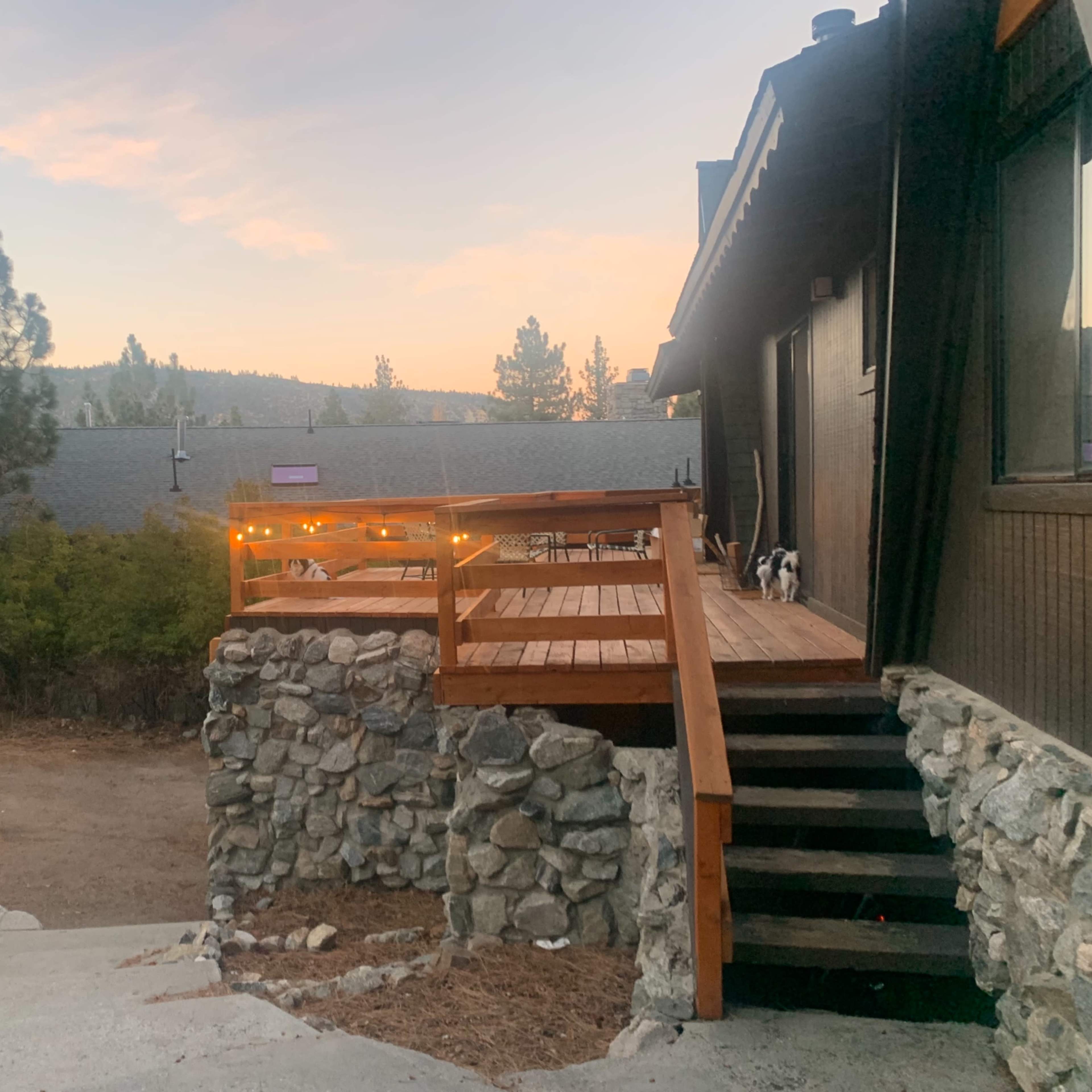 A wooden deck leading to a house, with stone steps and a view of trees and mountains in the background during sunset.