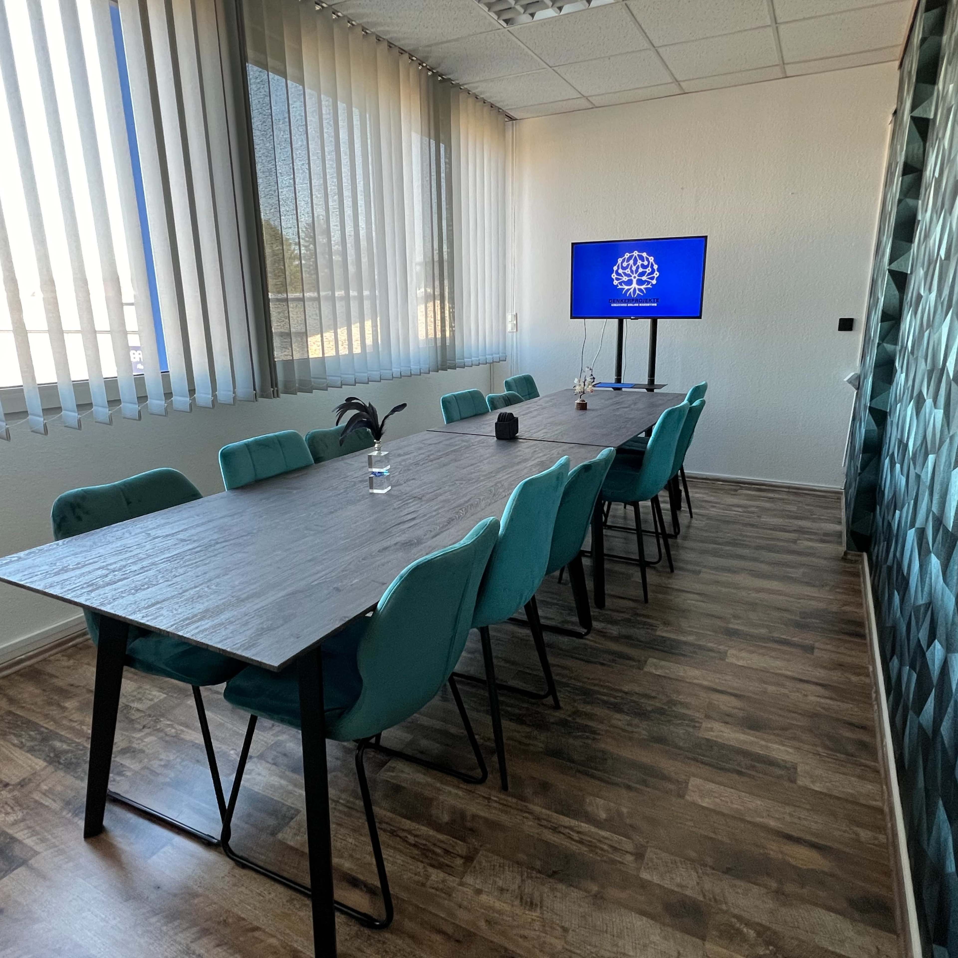 A conference room features a long table surrounded by teal chairs, with a television displaying a logo, and large windows covered by blinds.