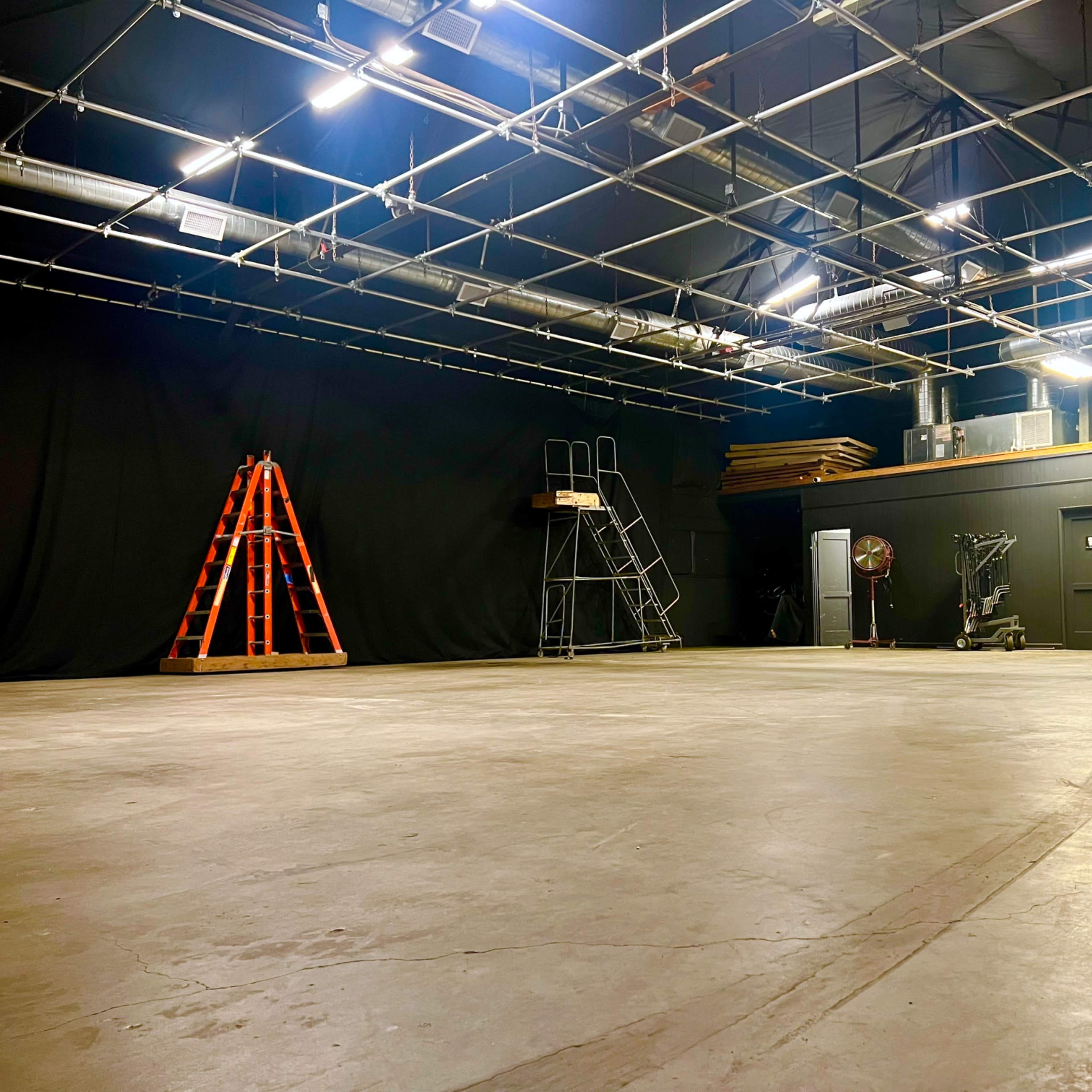 A dimly lit studio space with a bare concrete floor, black curtains lining the walls, and various equipment including a ladder, scaffolding, and a fan.