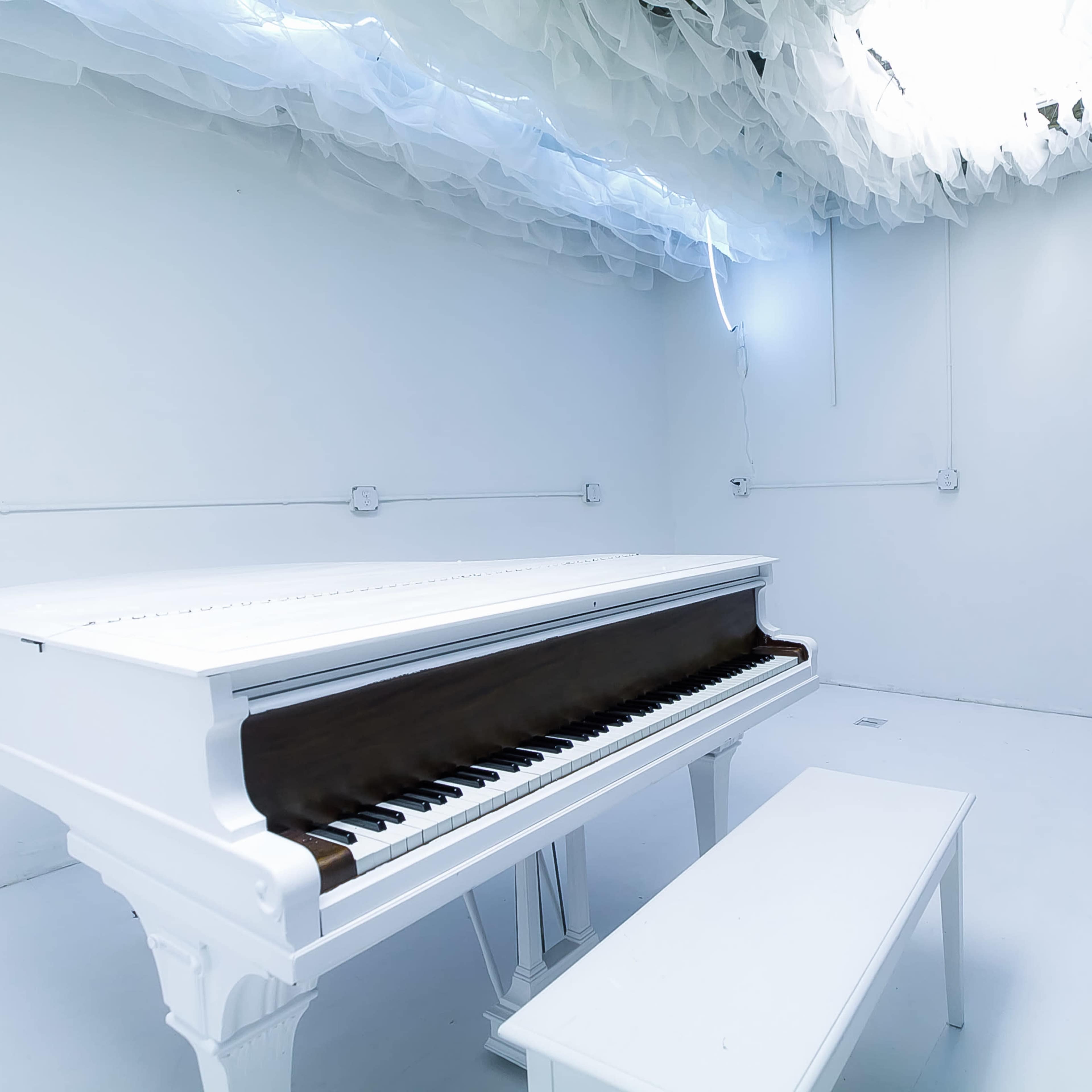 A white grand piano and matching bench are positioned in a minimalist, brightly lit room with a cloud-like ceiling decoration.