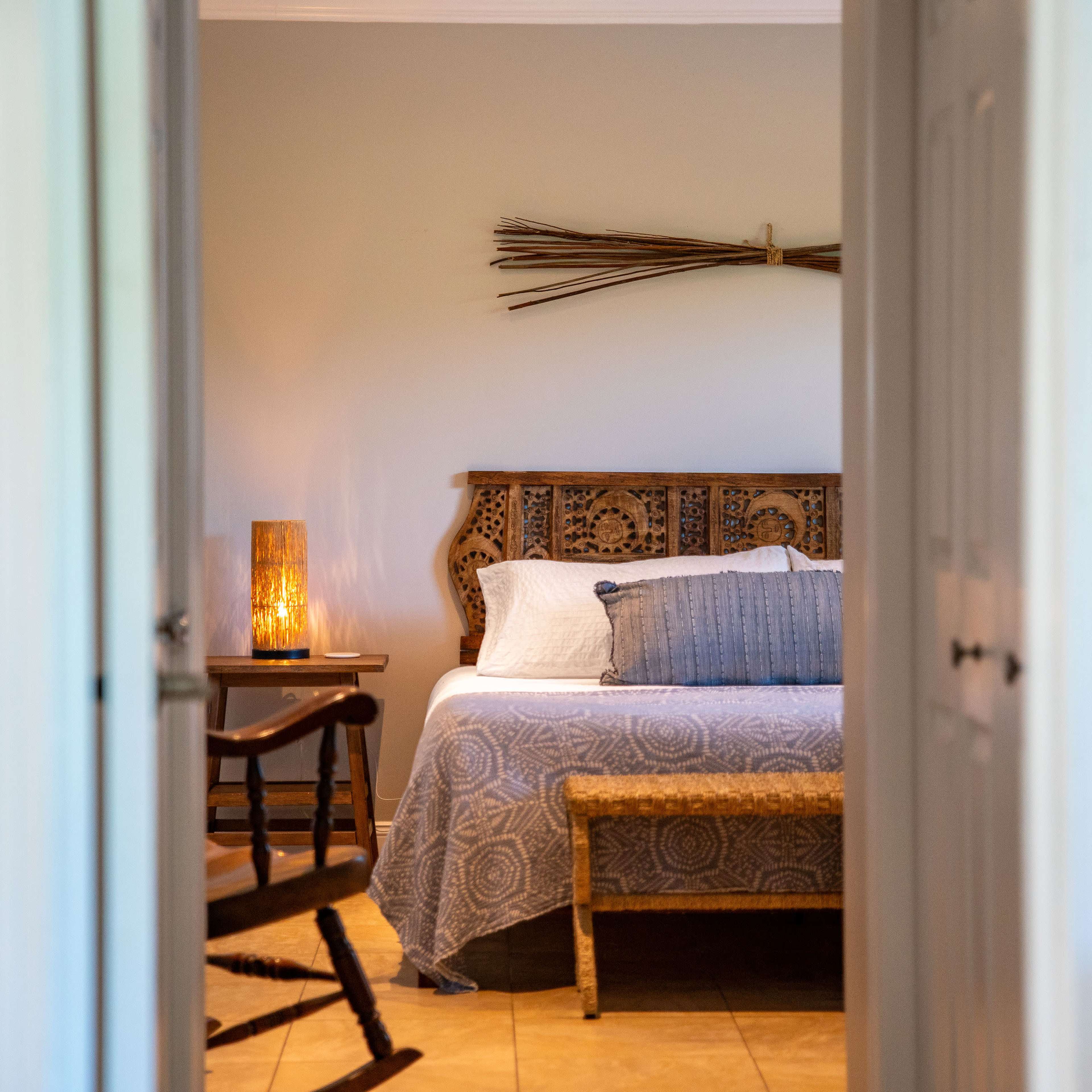 A neatly arranged bedroom features a wooden bed with a decorative headboard, a woven bench at the foot, and a lamp casting warm light.