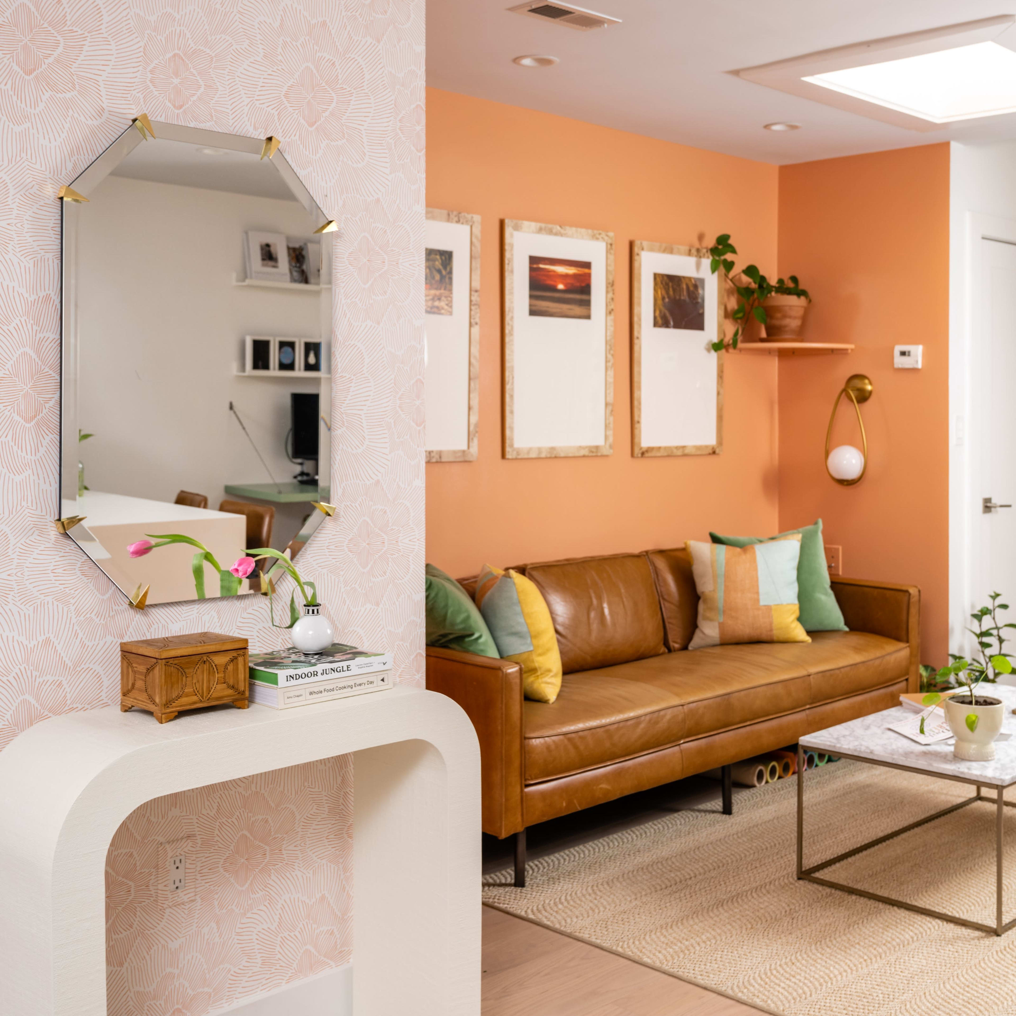 A living room with an orange wall, a brown leather sofa, a decorative mirror, and framed artwork hanging above a coffee table.
