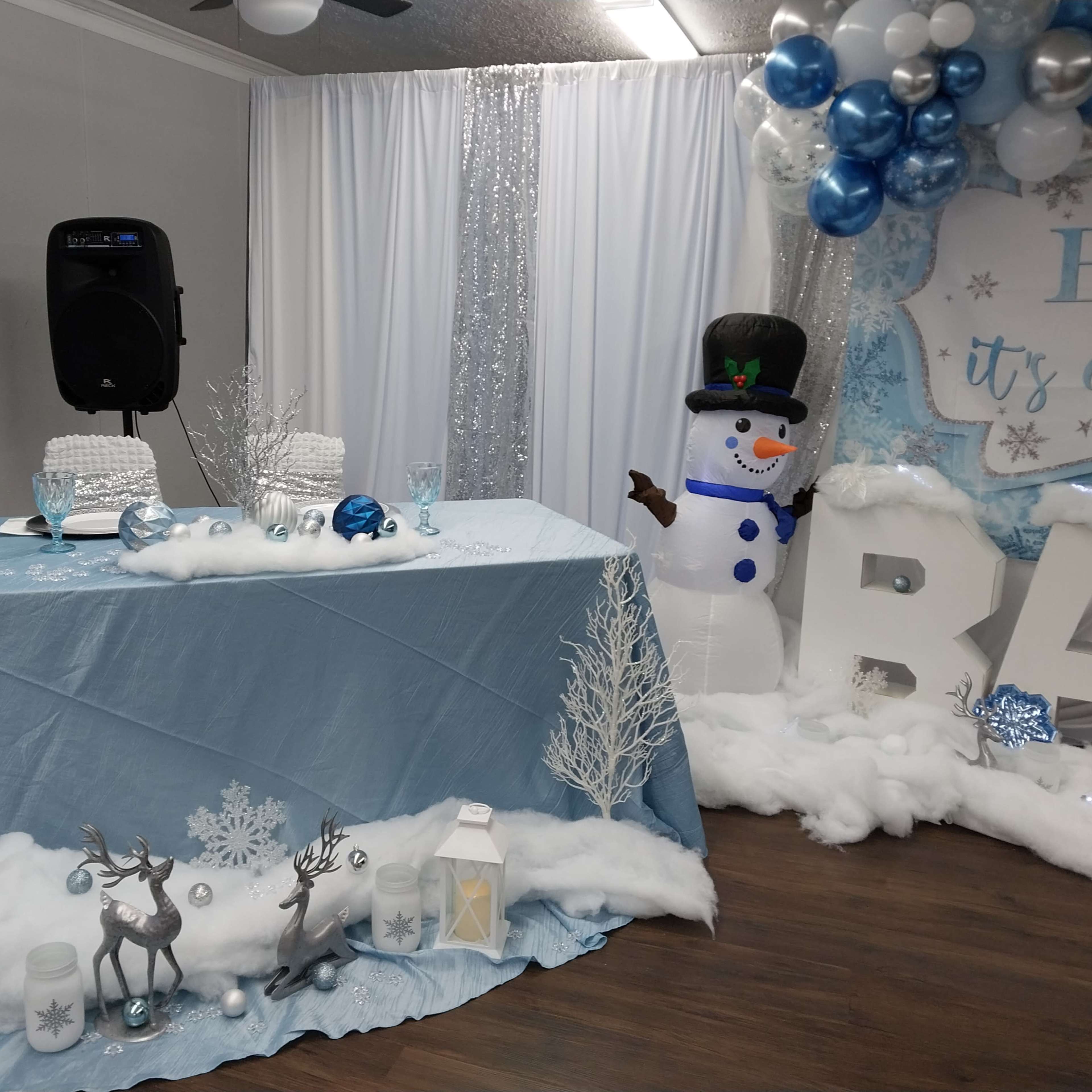 The scene features a decorated table with a blue tablecloth, a speaker, snowman, and winter-themed decorations including reindeer, lanterns, and balloons.