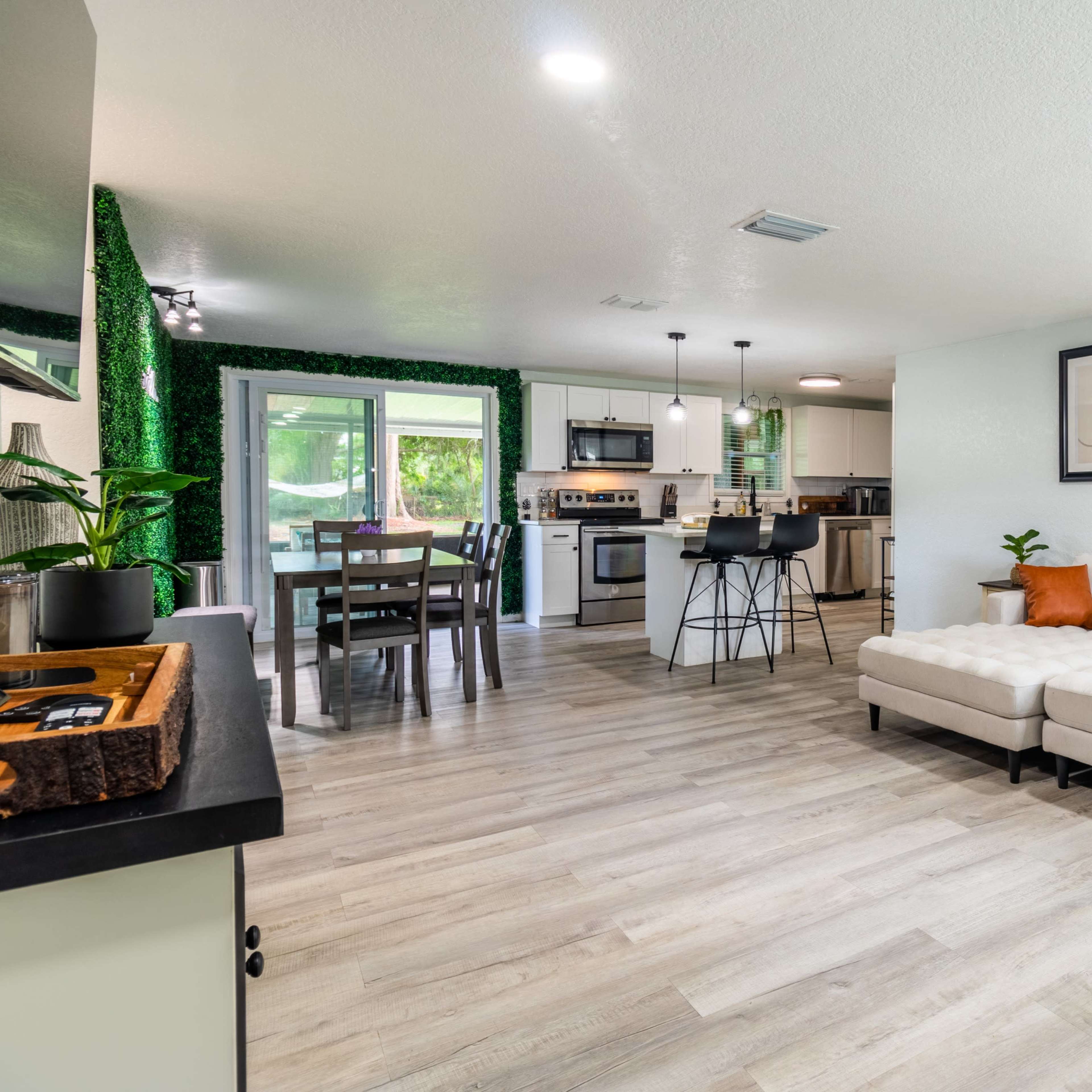 A modern open-concept living space featuring a white sectional sofa, a dining area with a wooden table, and a kitchen with stainless steel appliances.