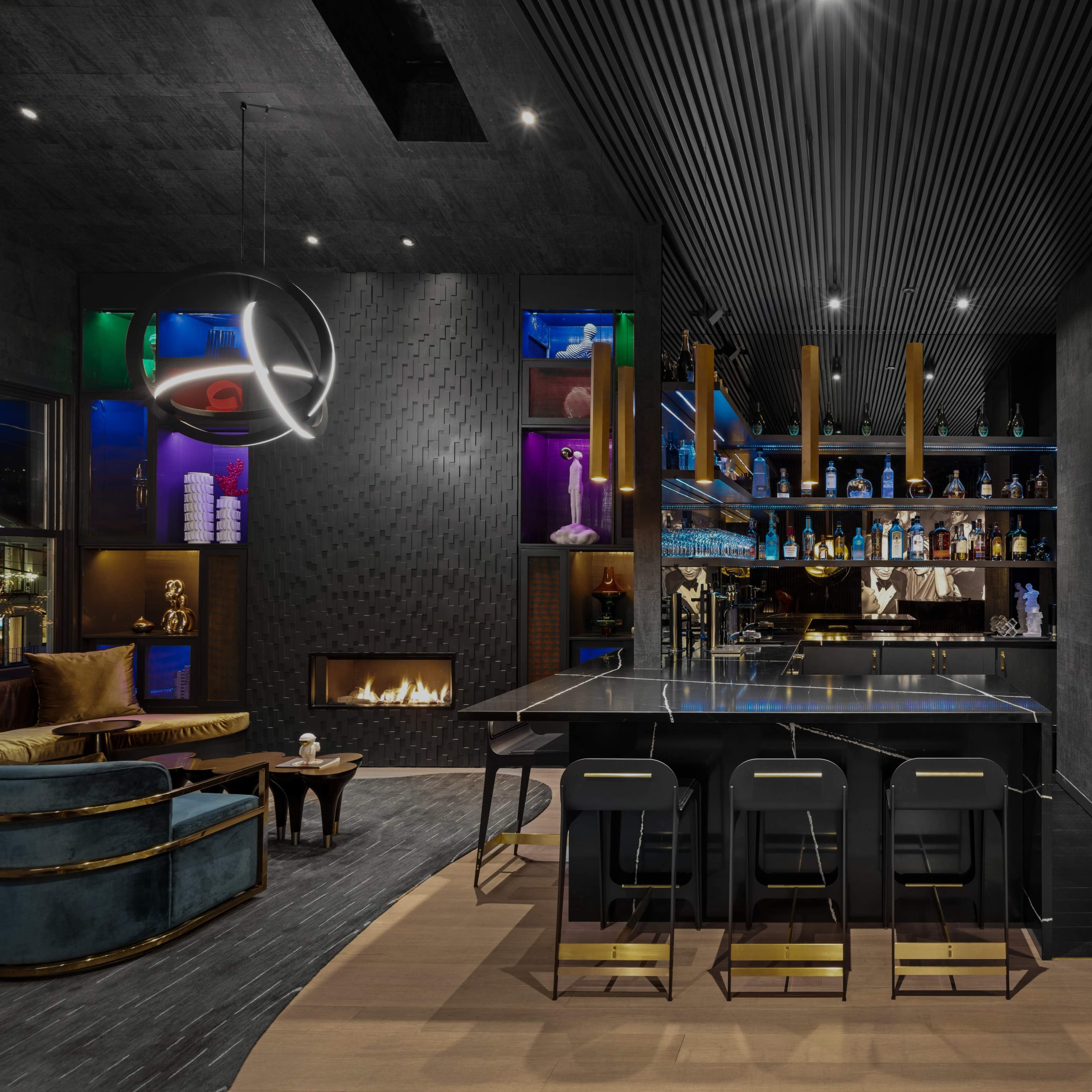 A modern bar area features a sleek black countertop, illuminated shelves displaying various bottles, and a cozy seating arrangement with a fireplace.