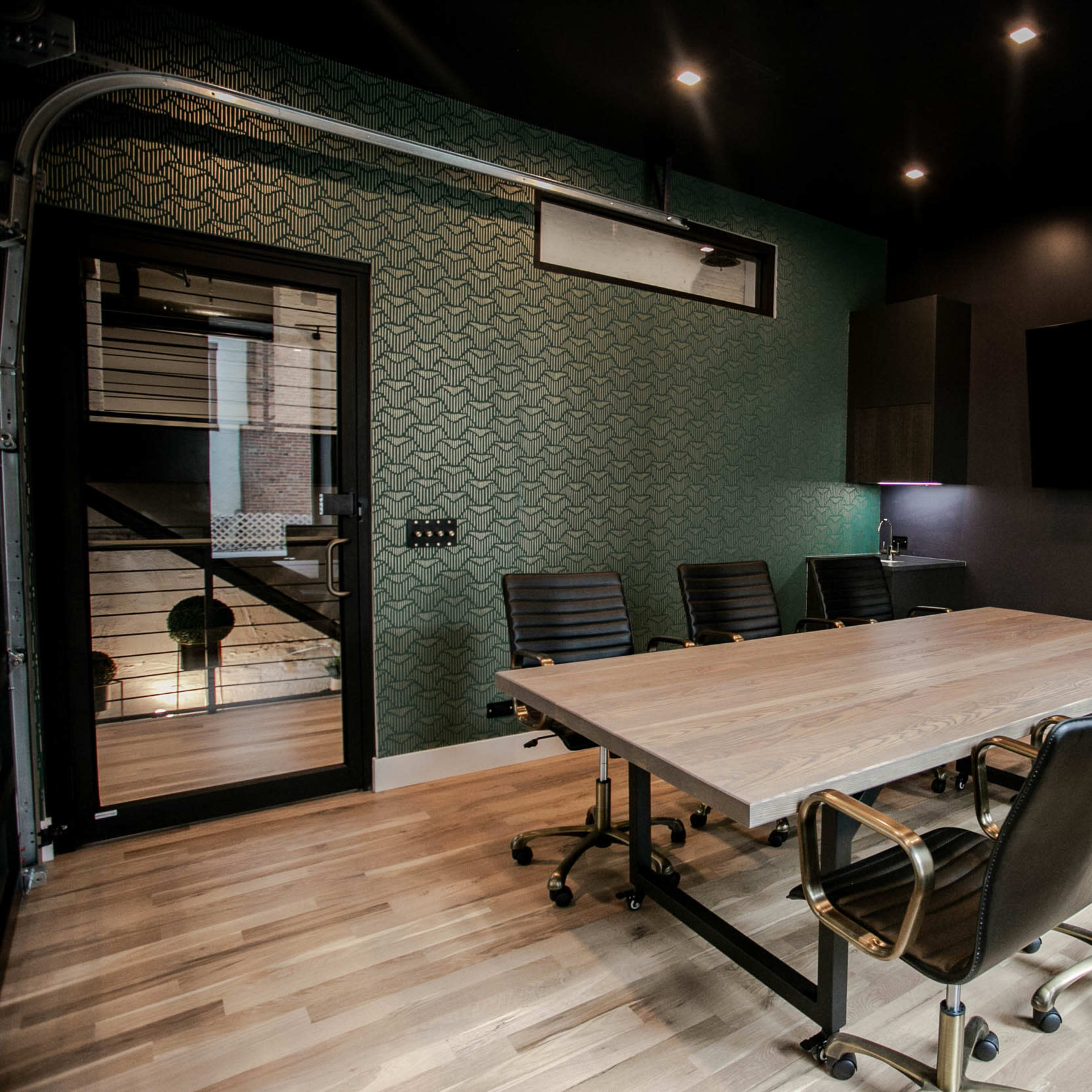 A modern conference room features a large wooden table, black ergonomic chairs, and a glass door leading to a staircase.