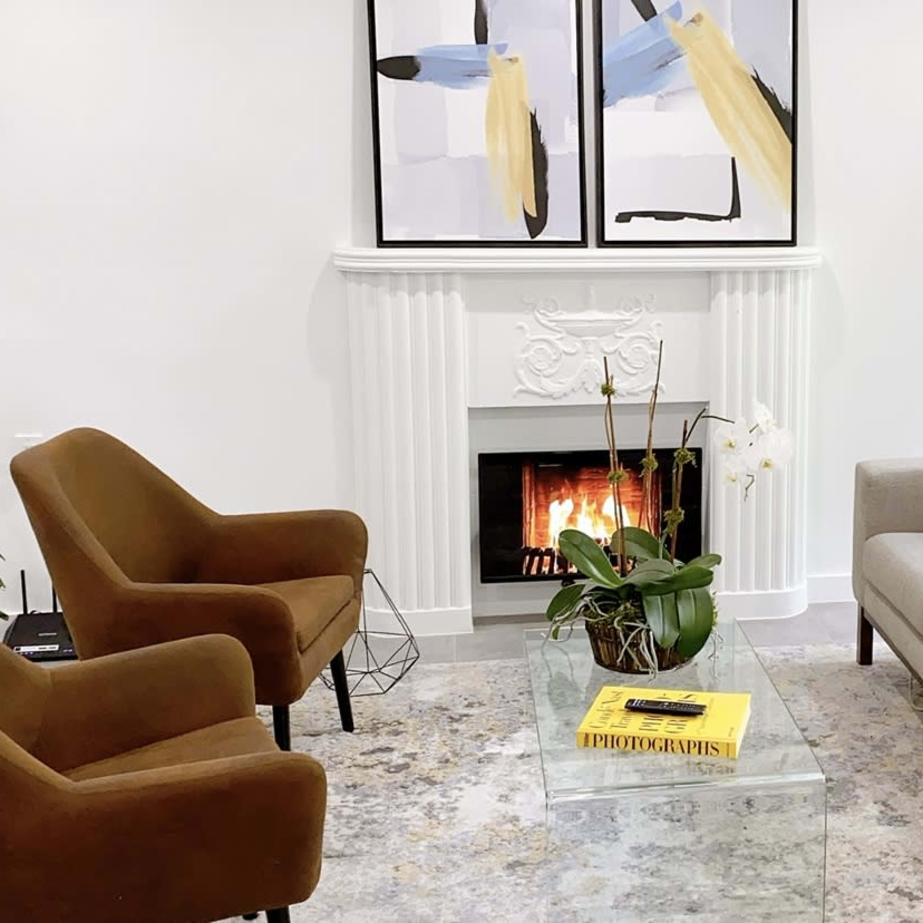 A modern living room with a beige sofa, two brown armchairs, a glass coffee table, a decorative plant, and a fireplace with a mantel, adorned with abstract art.