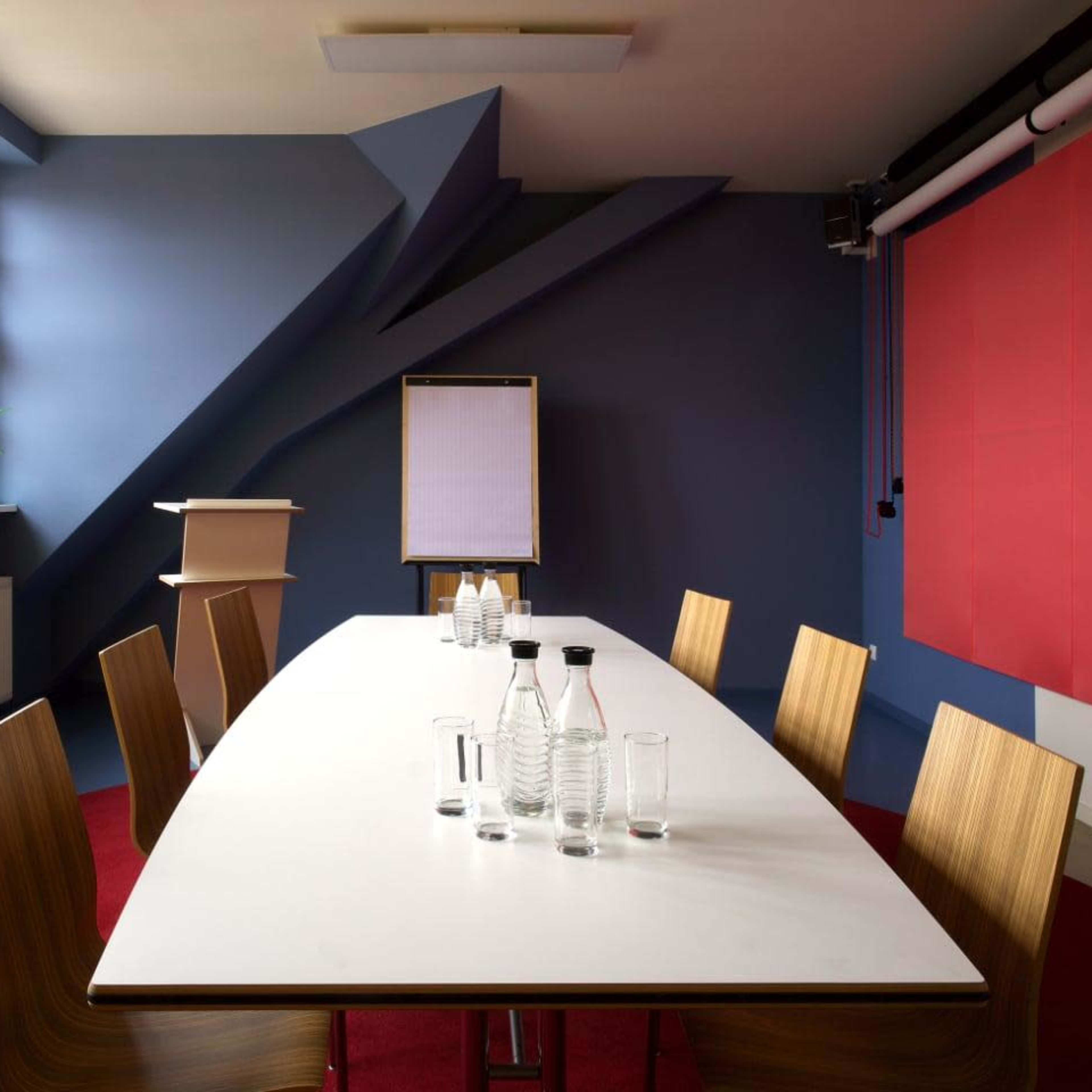 A modern conference room features a long white table surrounded by wooden chairs, with a flip chart and glass water bottles positioned on the table against a backdrop of colorful walls and large windows.