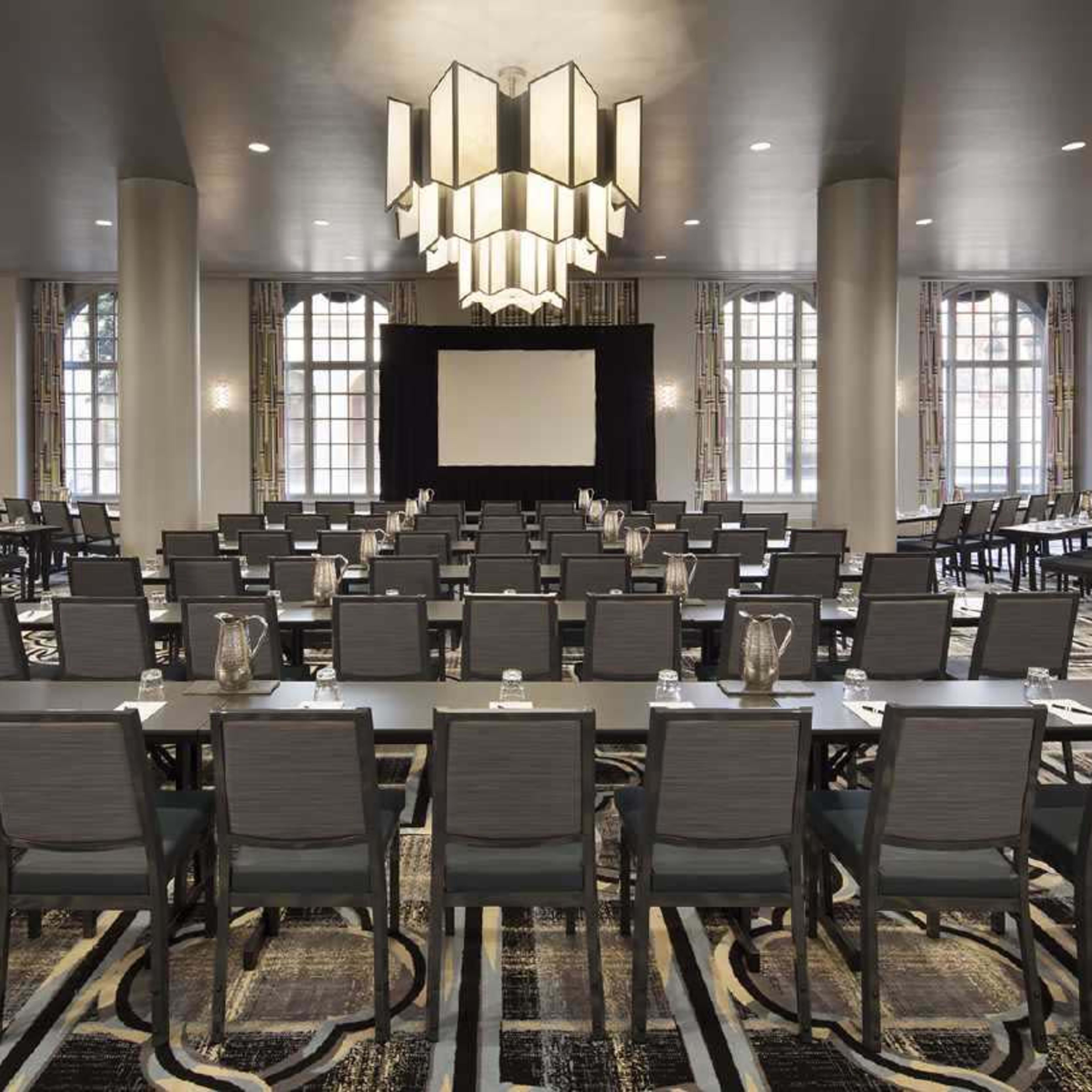 A conference room is arranged with rows of tables and chairs facing a large screen, illuminated by a modern chandelier.