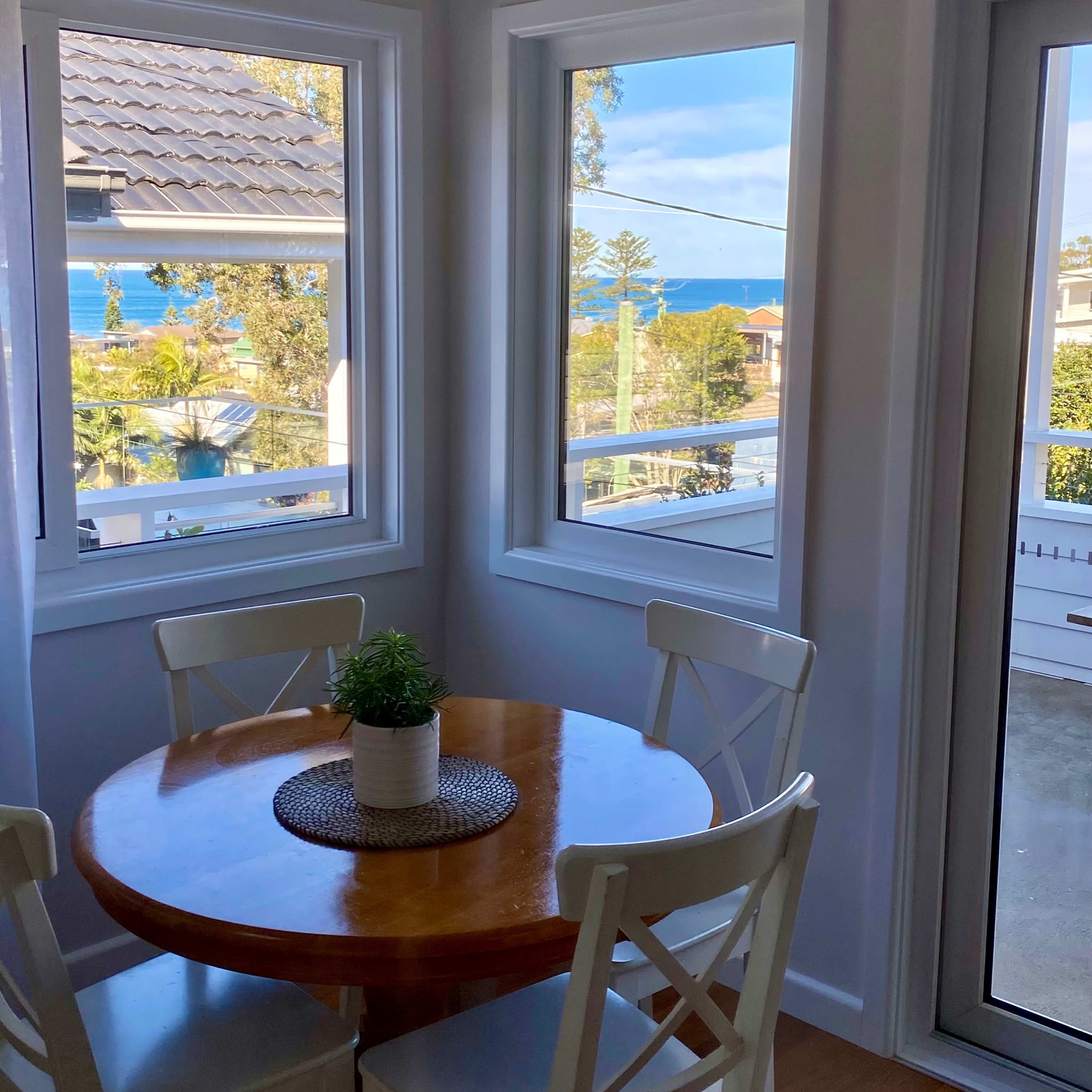 A round wooden table with four white chairs is set against a backdrop of large windows overlooking a coastal view.