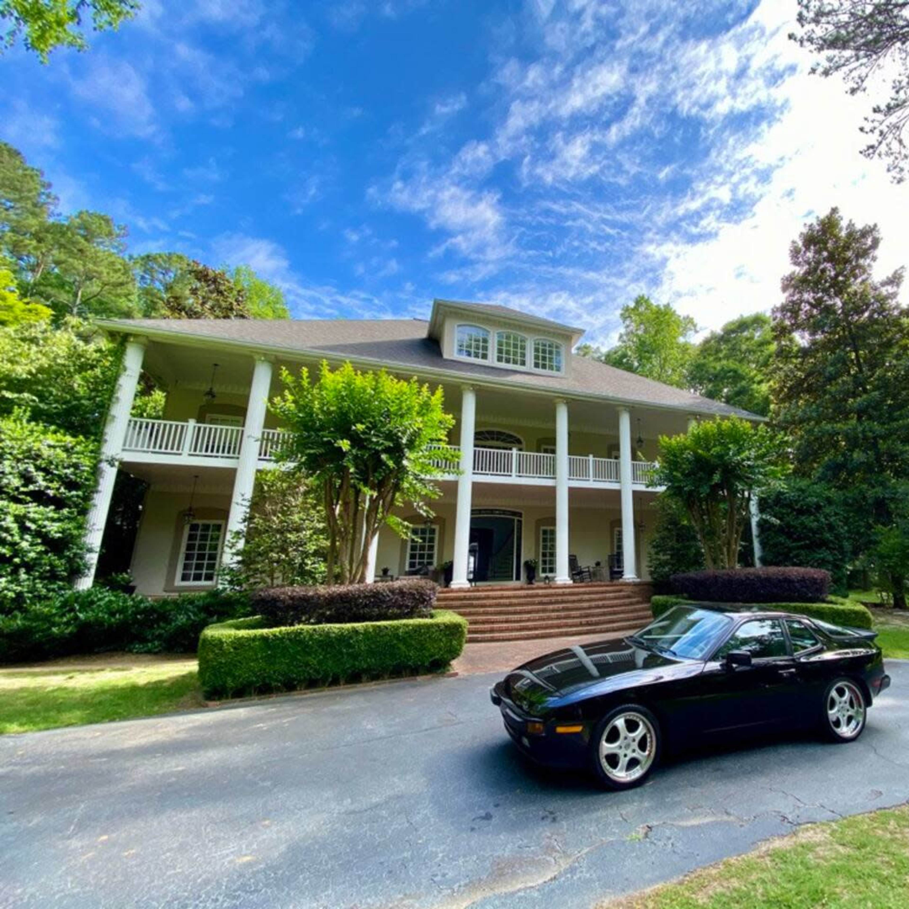 A black sports car is parked in front of a large two-story house surrounded by greenery.