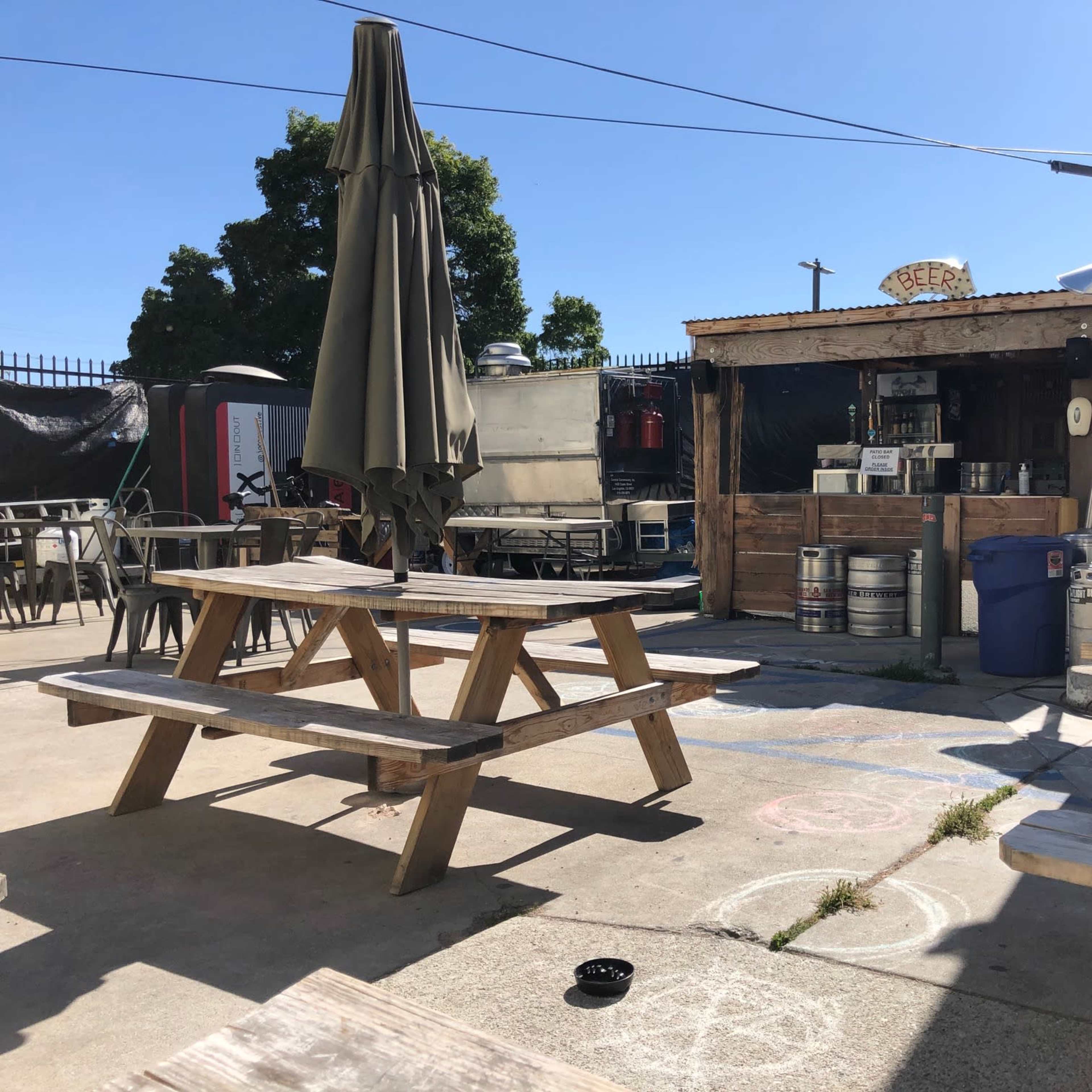 An outdoor seating area with wooden picnic tables in front of a small building that appears to serve drinks, including beer kegs and a television mounted on the wall.