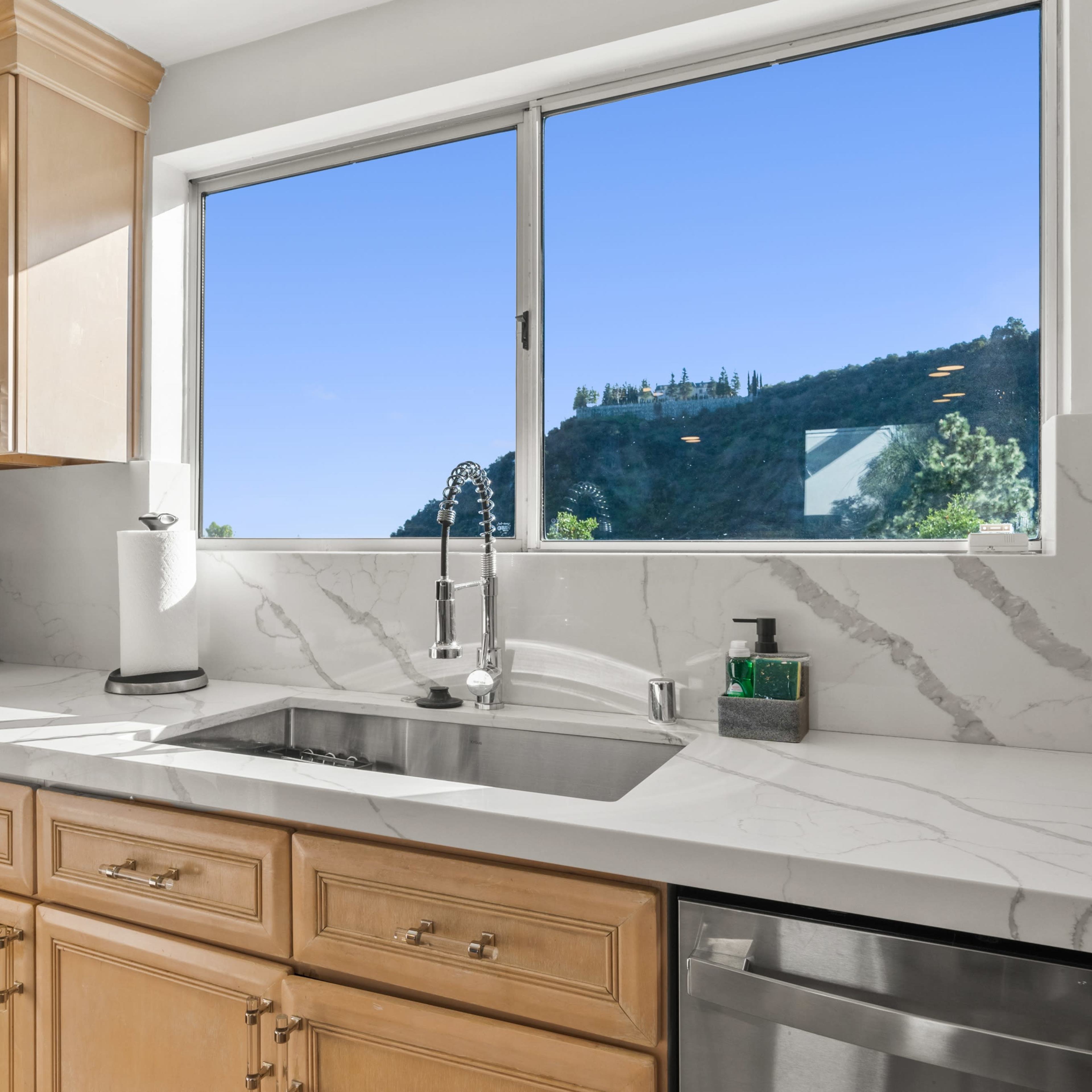 A bright kitchen with light wooden cabinets, a marble countertop, and a window overlooking a green hillside.