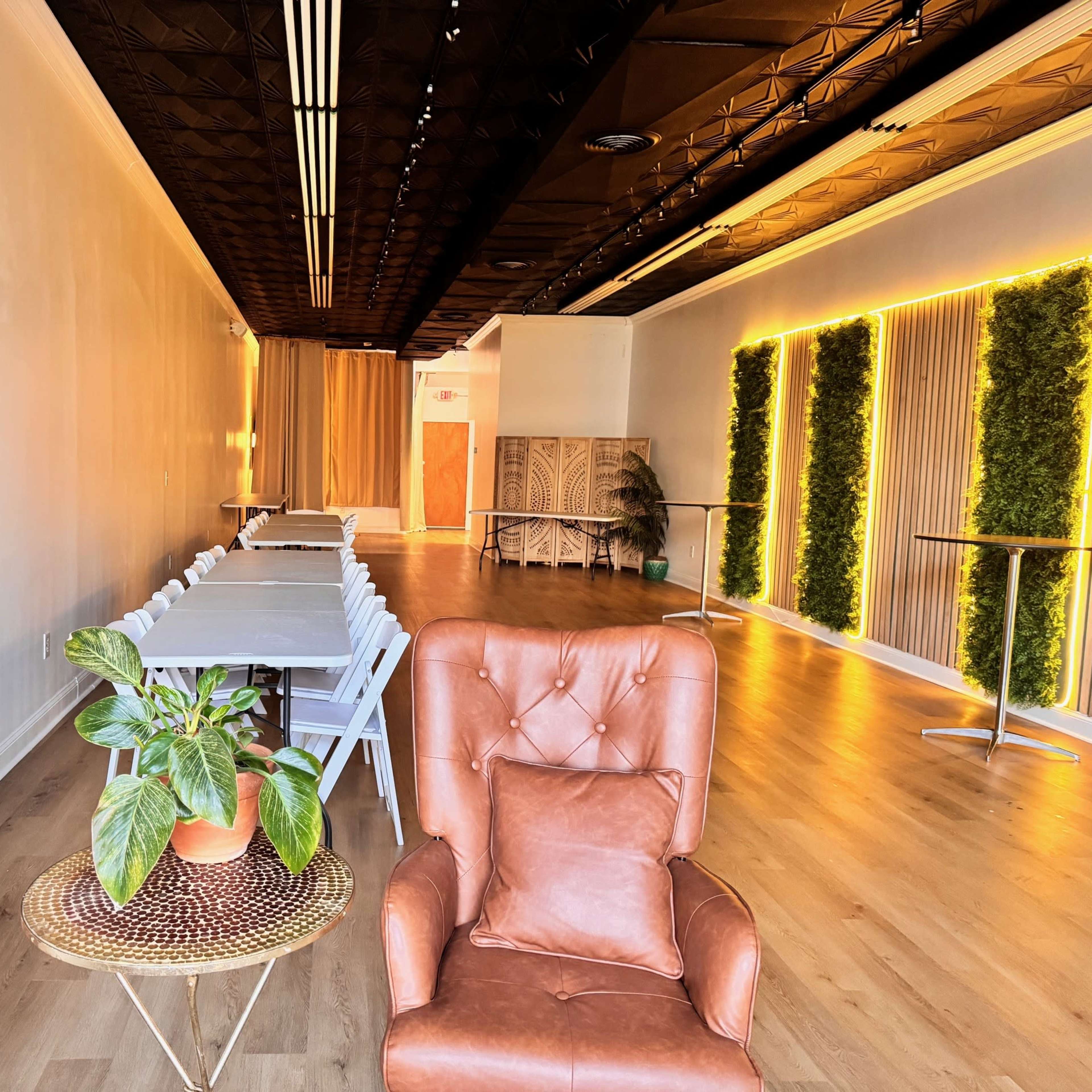 A spacious interior with brown armchairs, a row of tables and chairs, and wall-mounted greenery lighting.