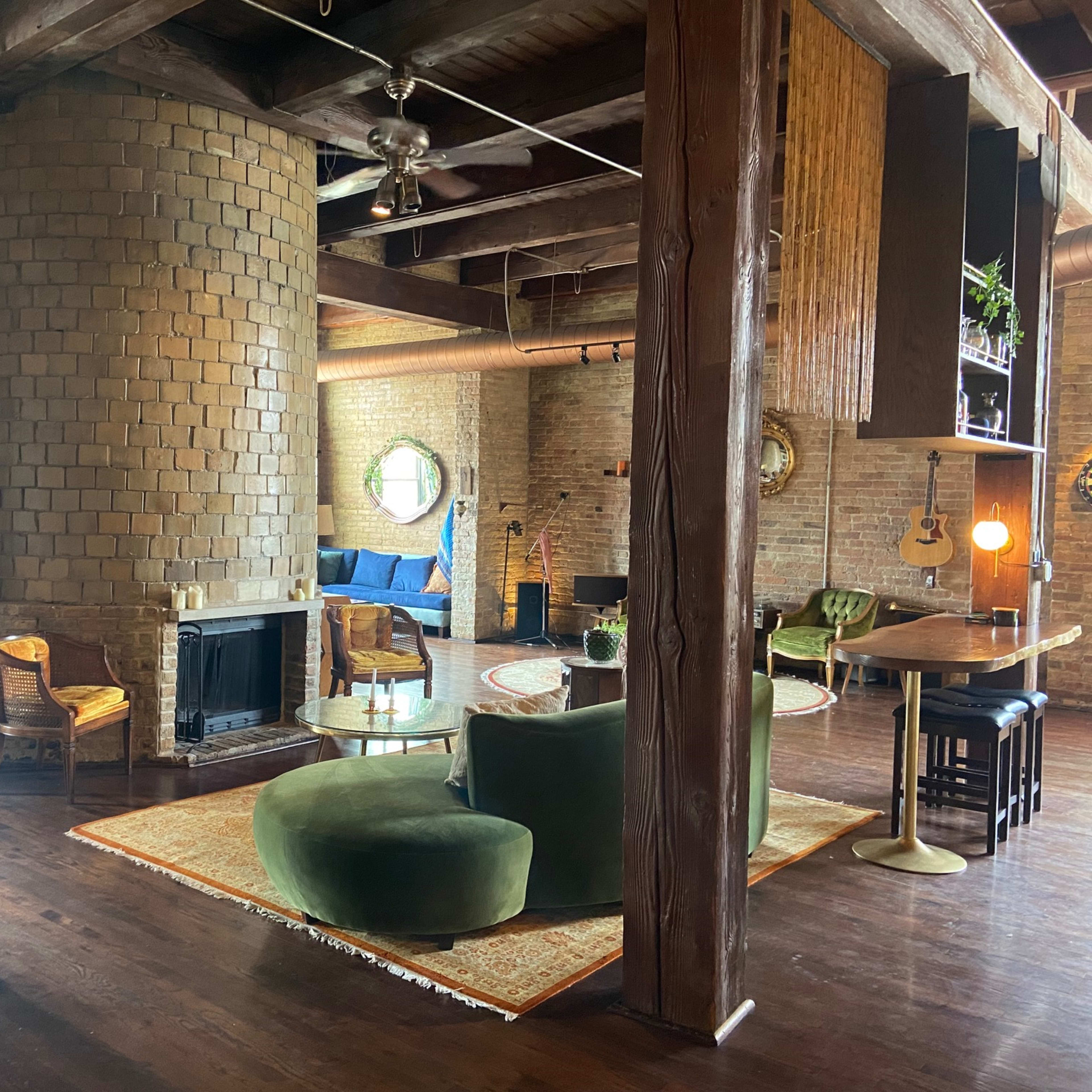 The interior features a spacious room with exposed brick walls, wooden beams, and a circular fireplace, surrounded by various seating areas and decorative elements.