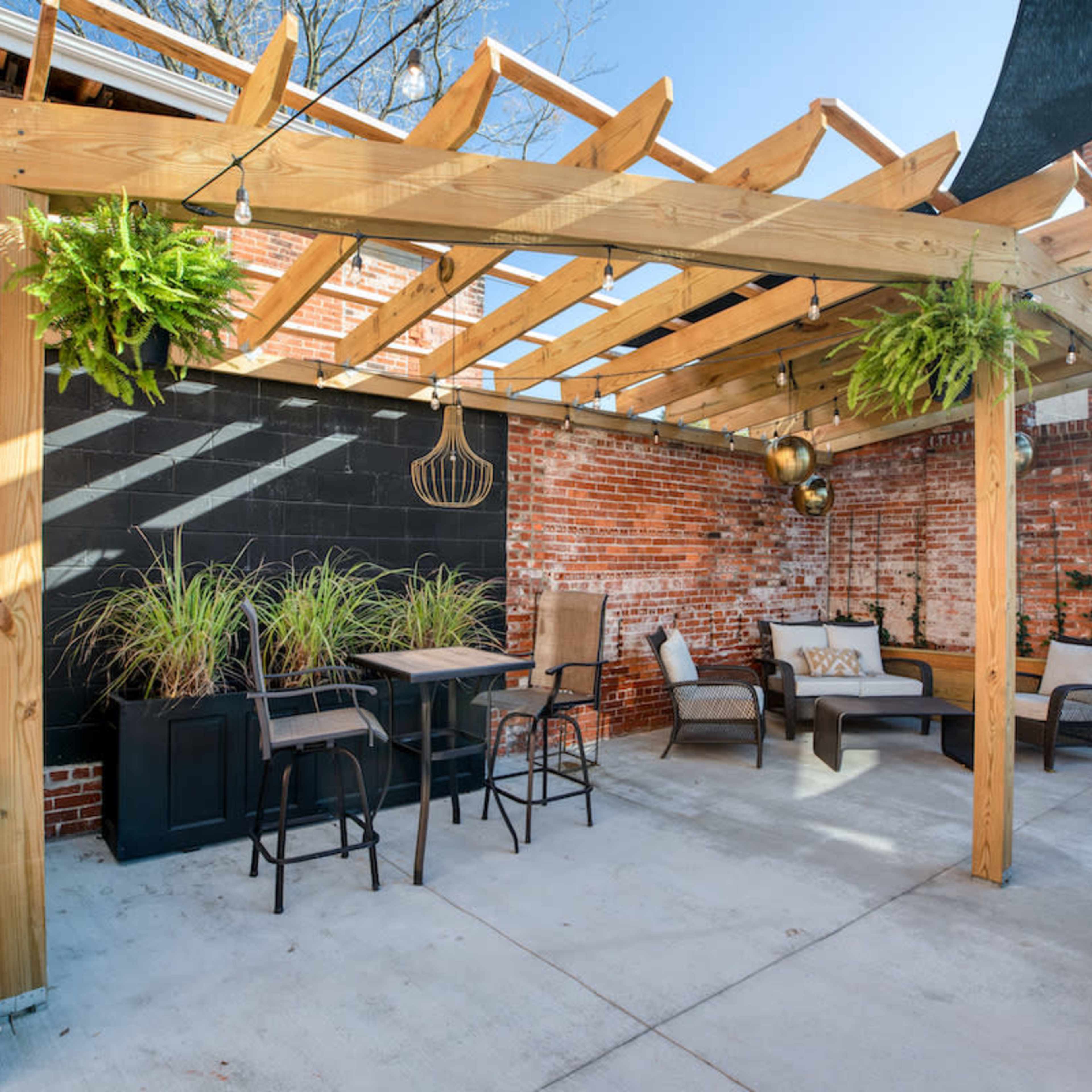 A patio area with wooden pergola, seating arrangements, and planters against a brick wall.