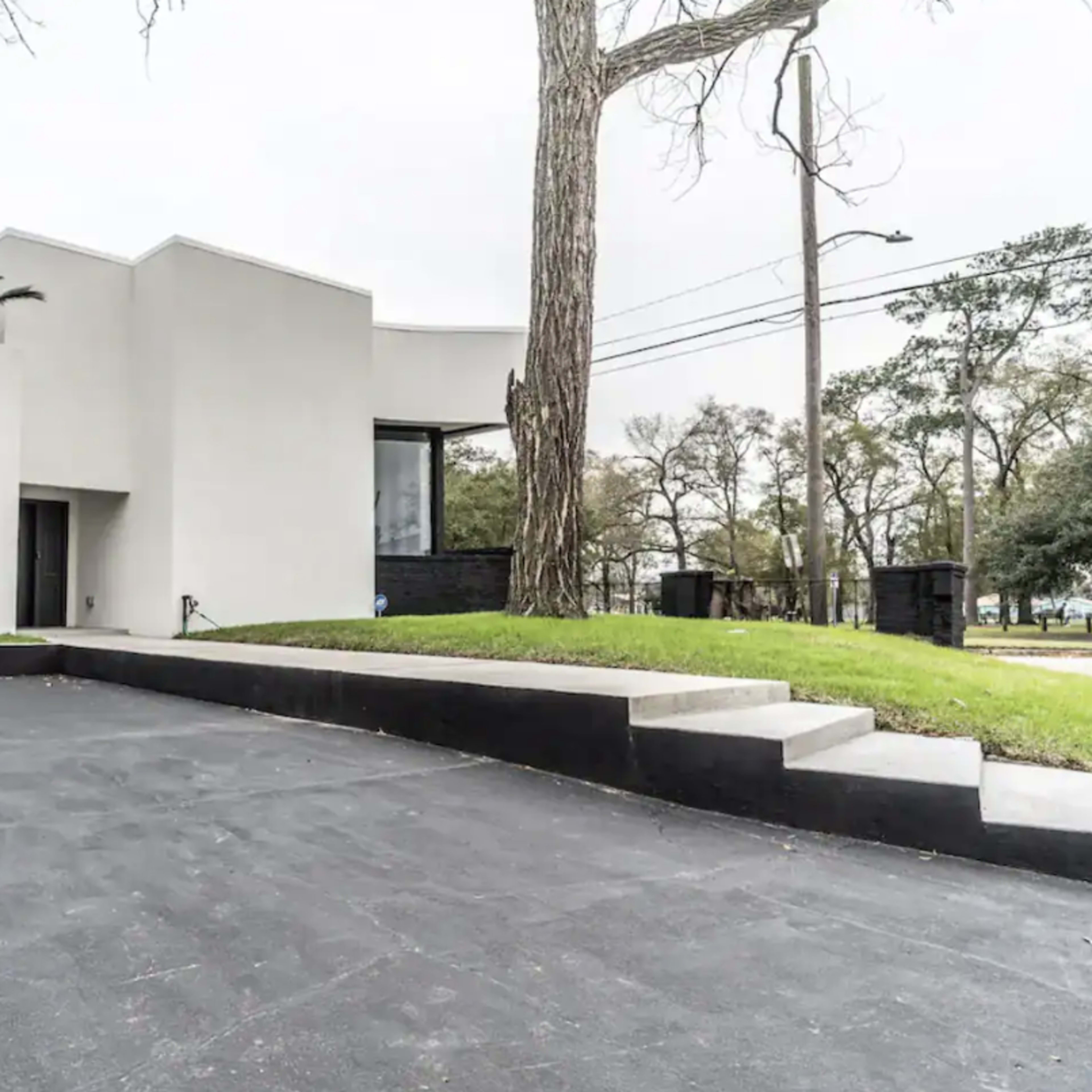 A modern, minimalist white house with large windows and a flat roof, surrounded by green grass and trees, with a concrete driveway leading to the entrance.