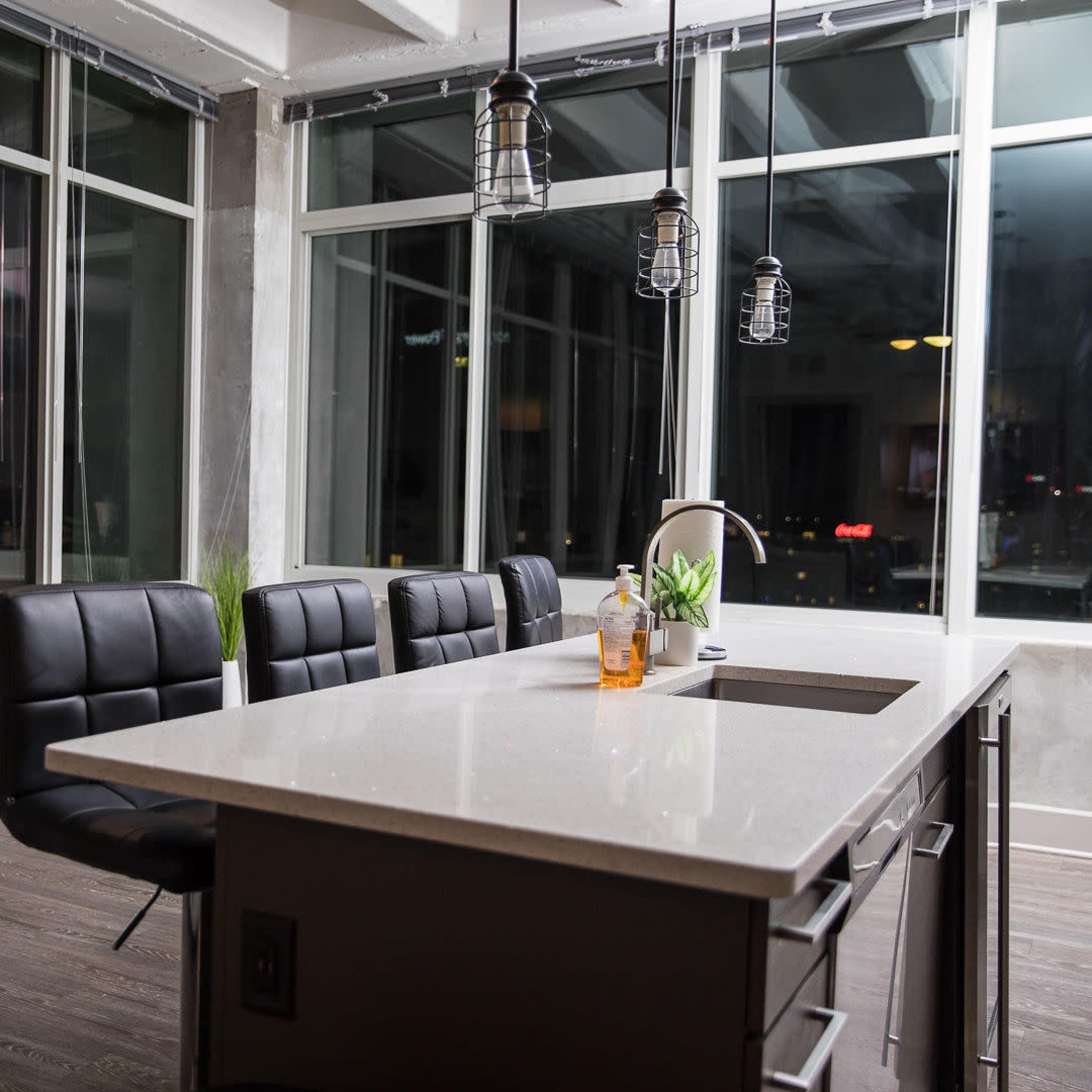 A modern kitchen features a central island with a white countertop, surrounded by black leather chairs and large windows offering a view of the city.