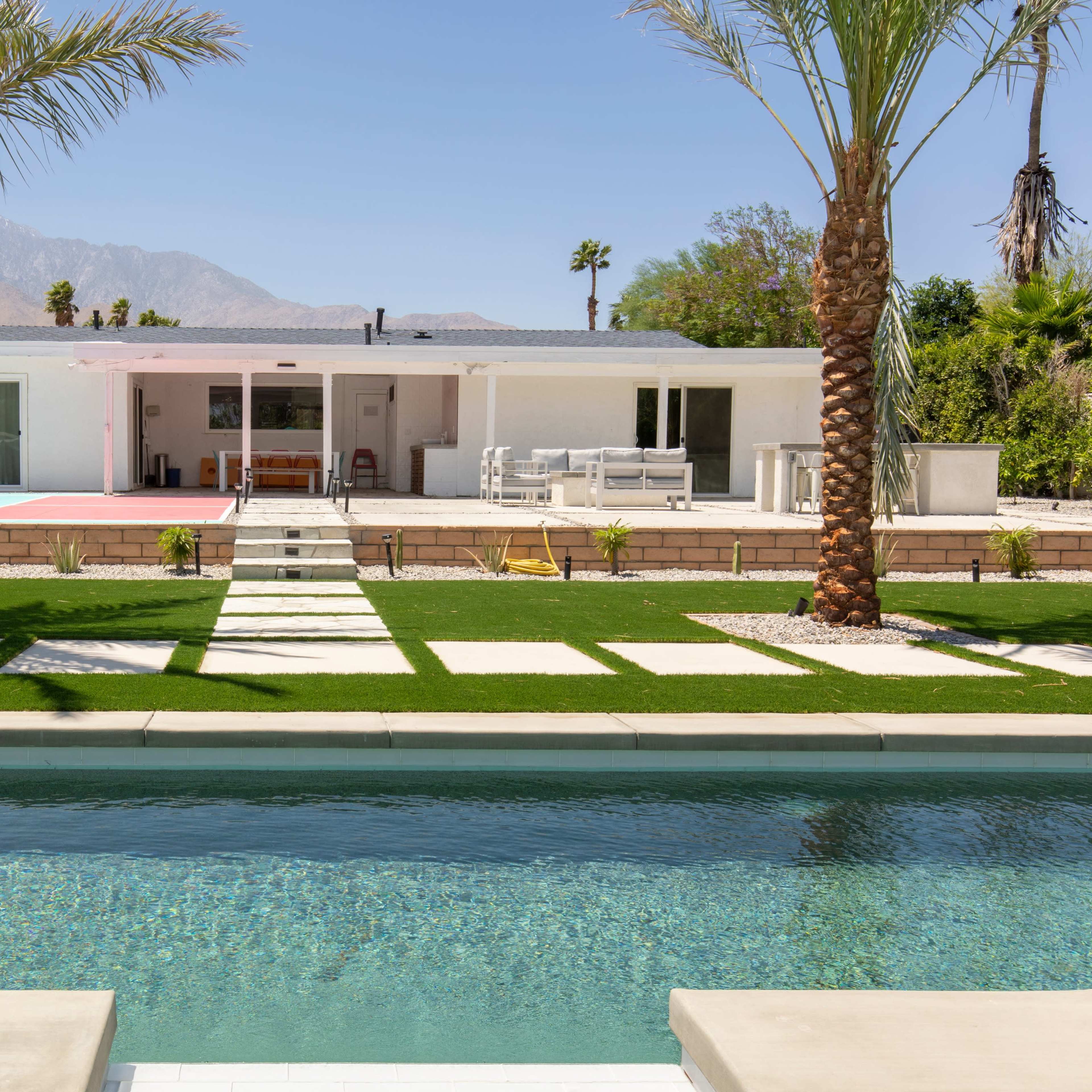 A modern white house with palm trees, a pool, and recreational areas in a desert landscape.