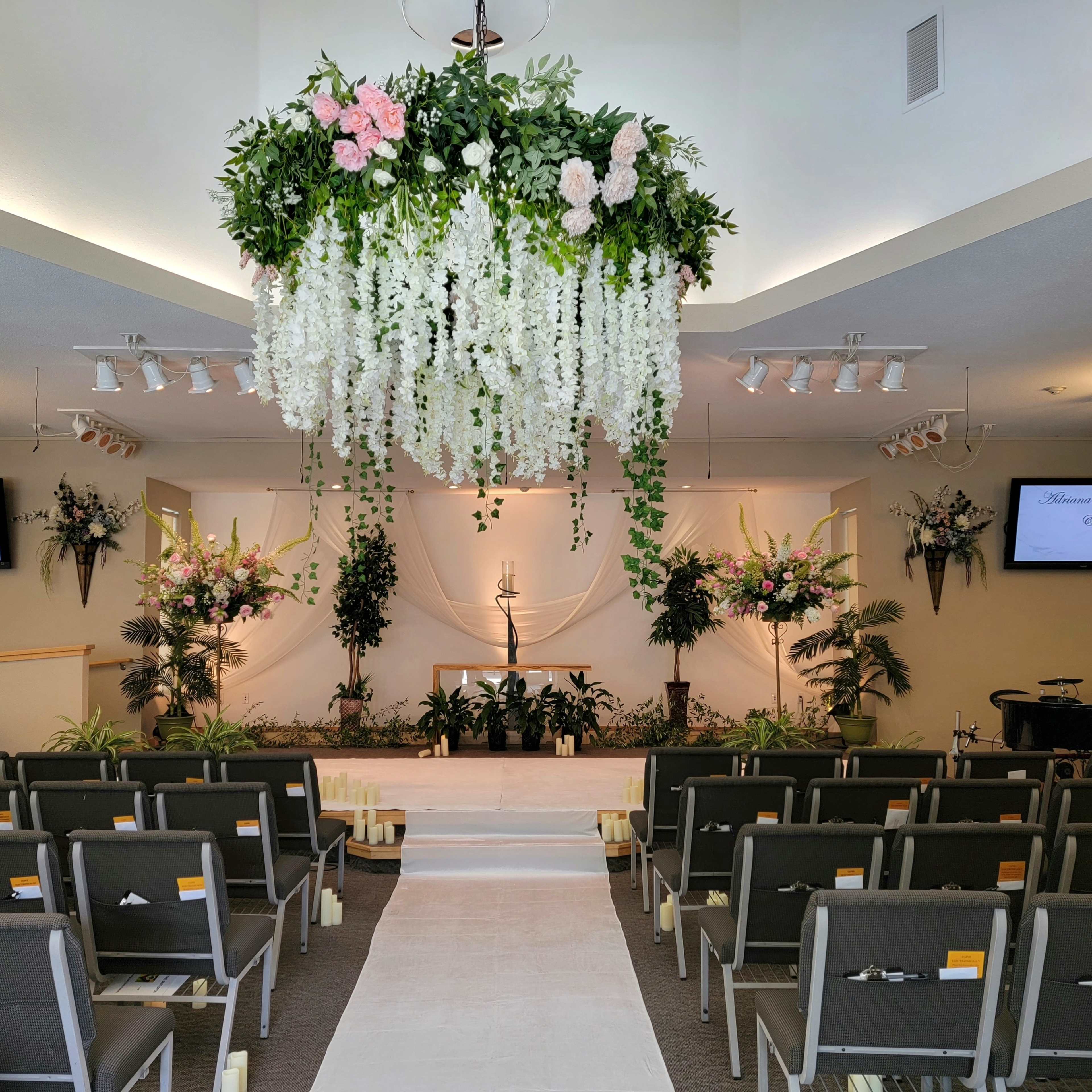 A beautifully arranged wedding venue features a floral canopy above an altar, flanked by decorative plants and two screens displaying welcoming messages.