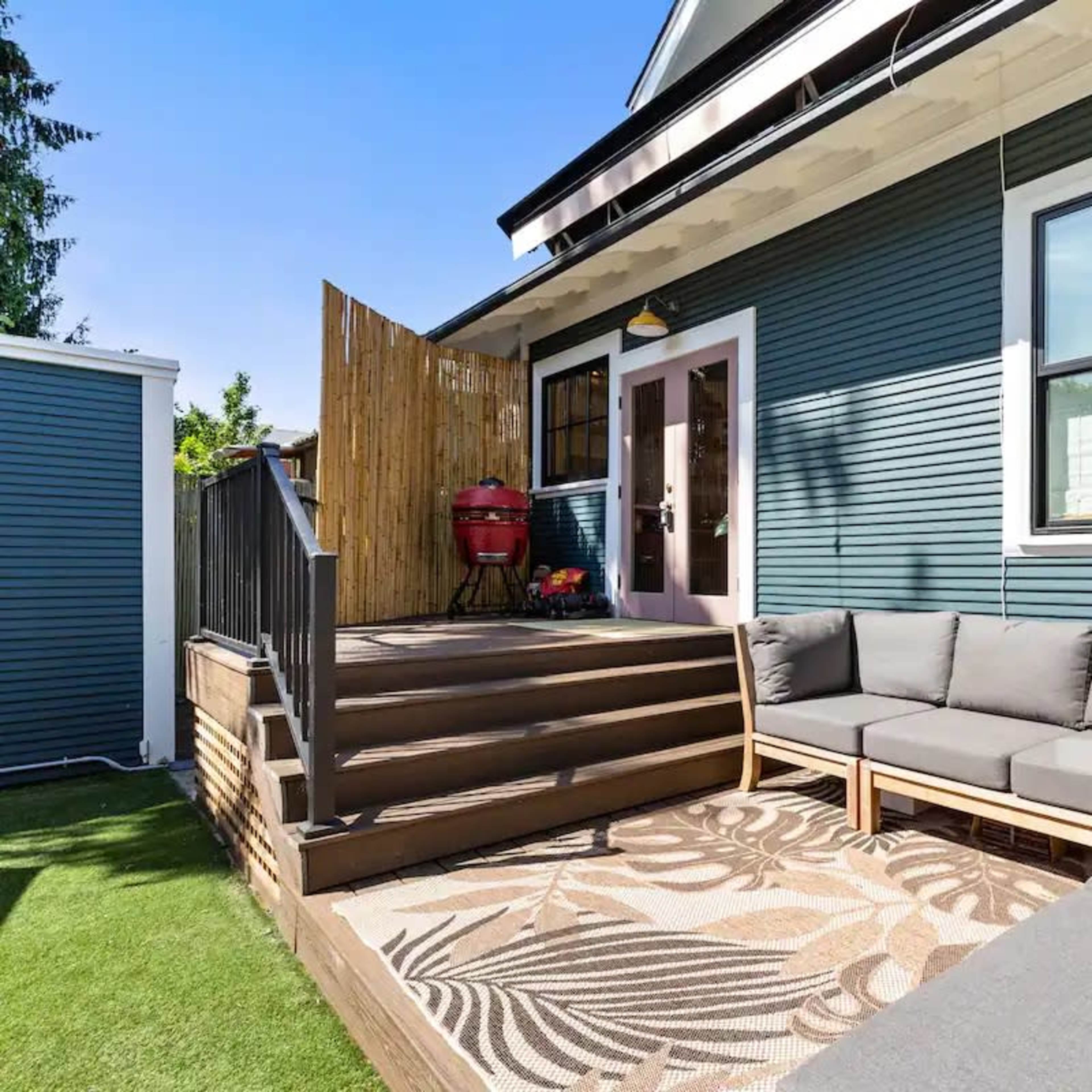 A house with a gray-blue exterior and a wooden deck featuring a sectional sofa and a decorative rug.