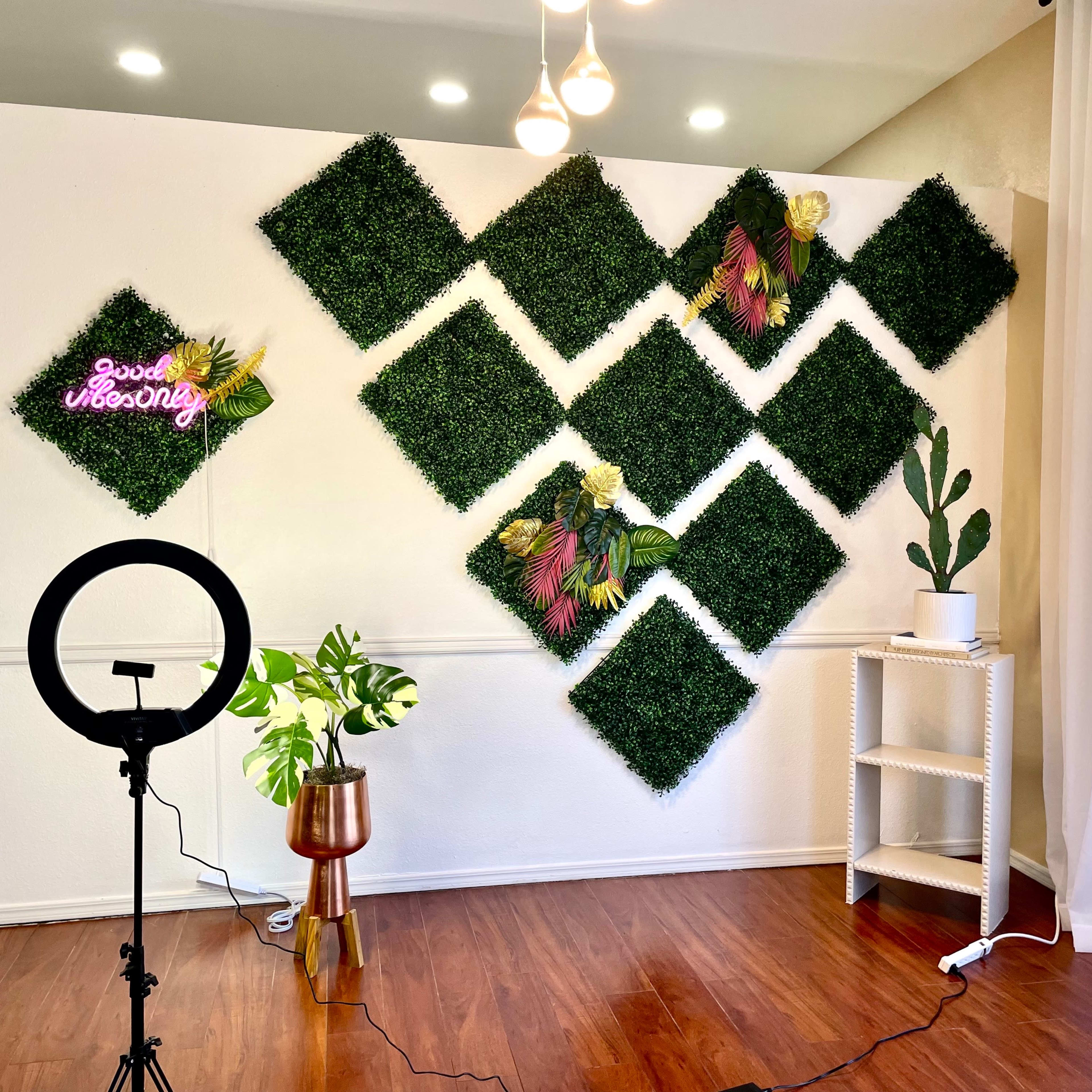 A room with a decorative wall of artificial greenery arranged in diamond shapes, complemented by a neon sign and potted plants.