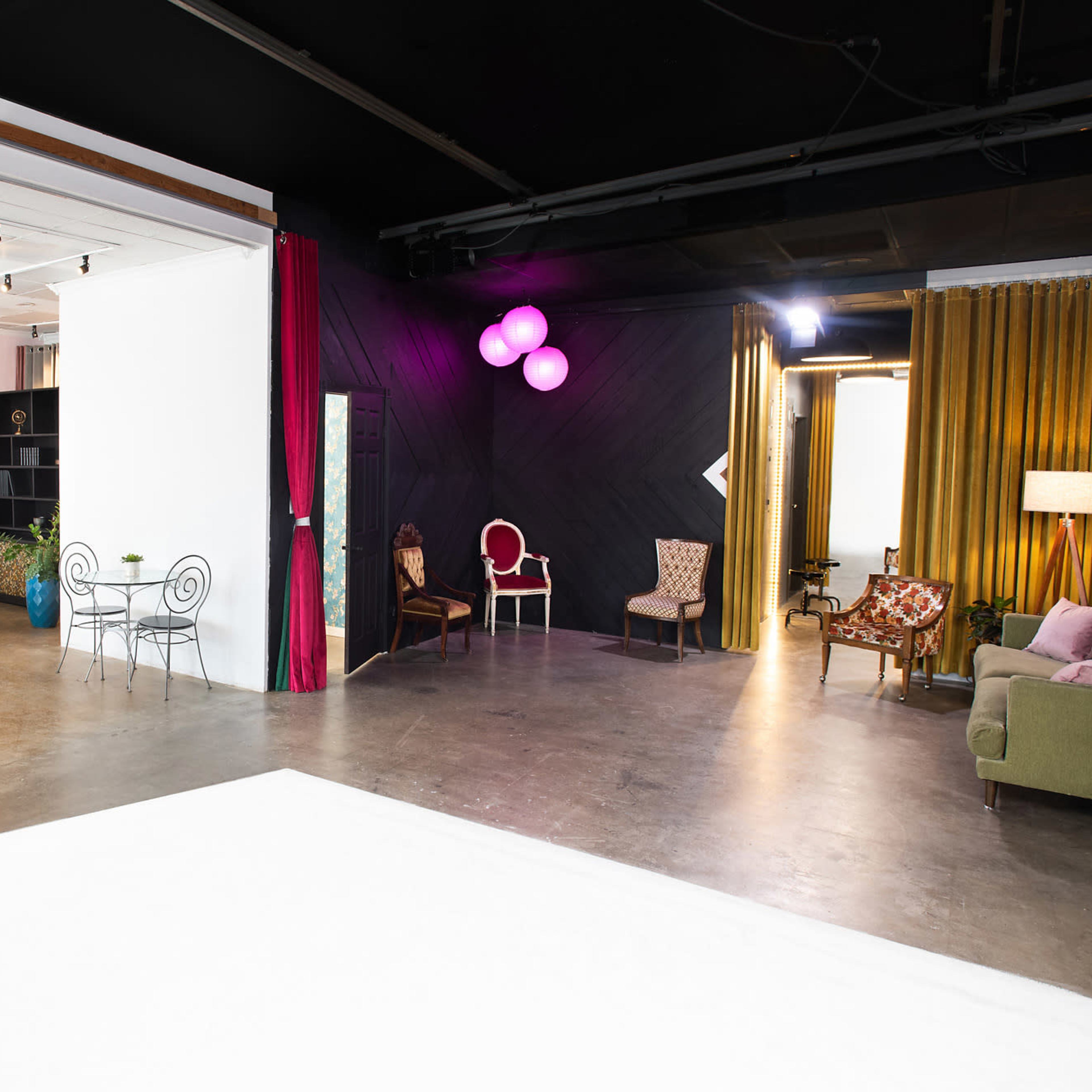 A spacious studio divided into two areas, featuring a dining setup with chairs and a table on one side and a cozy seating arrangement with plants and decorative curtains on the other.