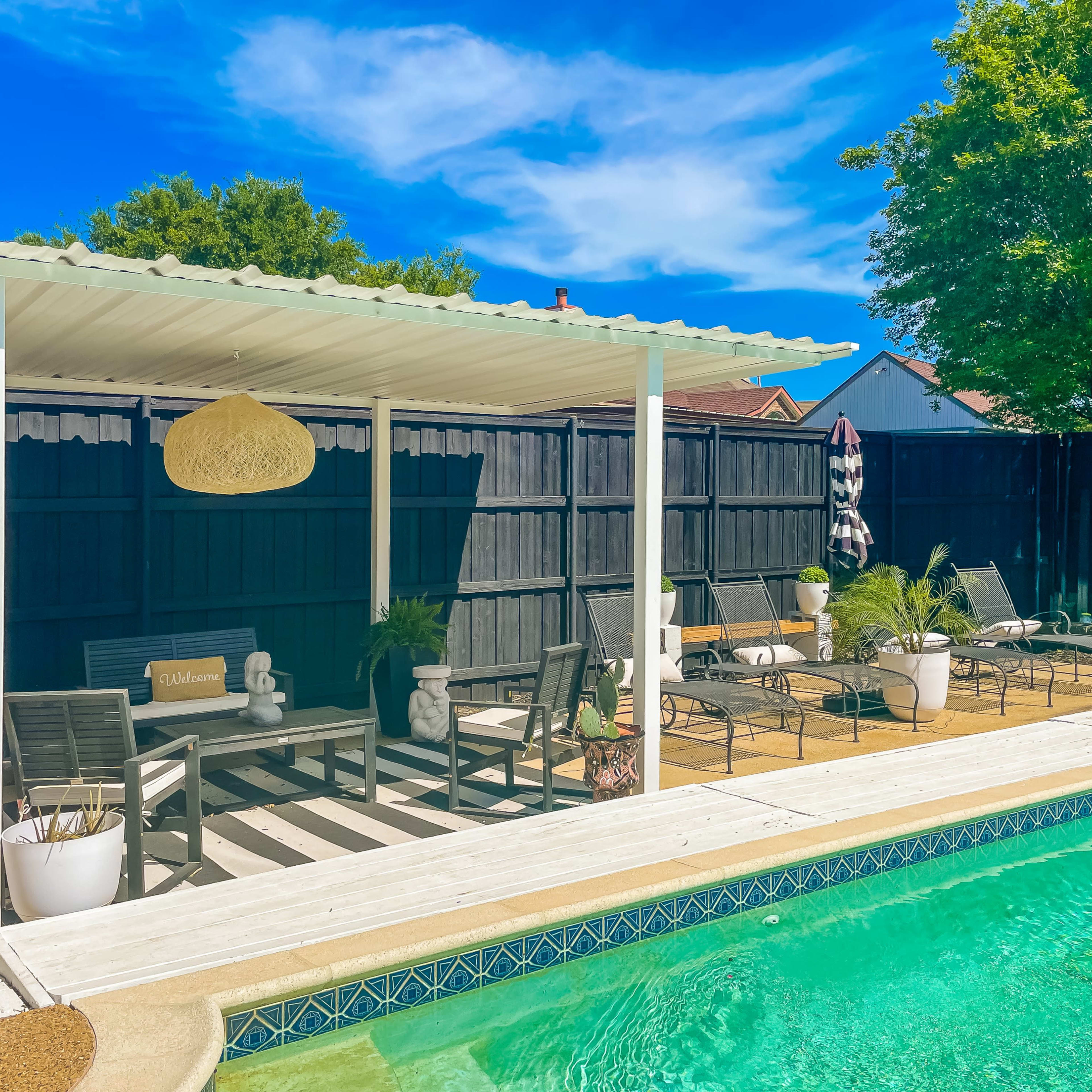 A swimming pool area with a covered seating space, lounge chairs, potted plants, and a clear blue sky.