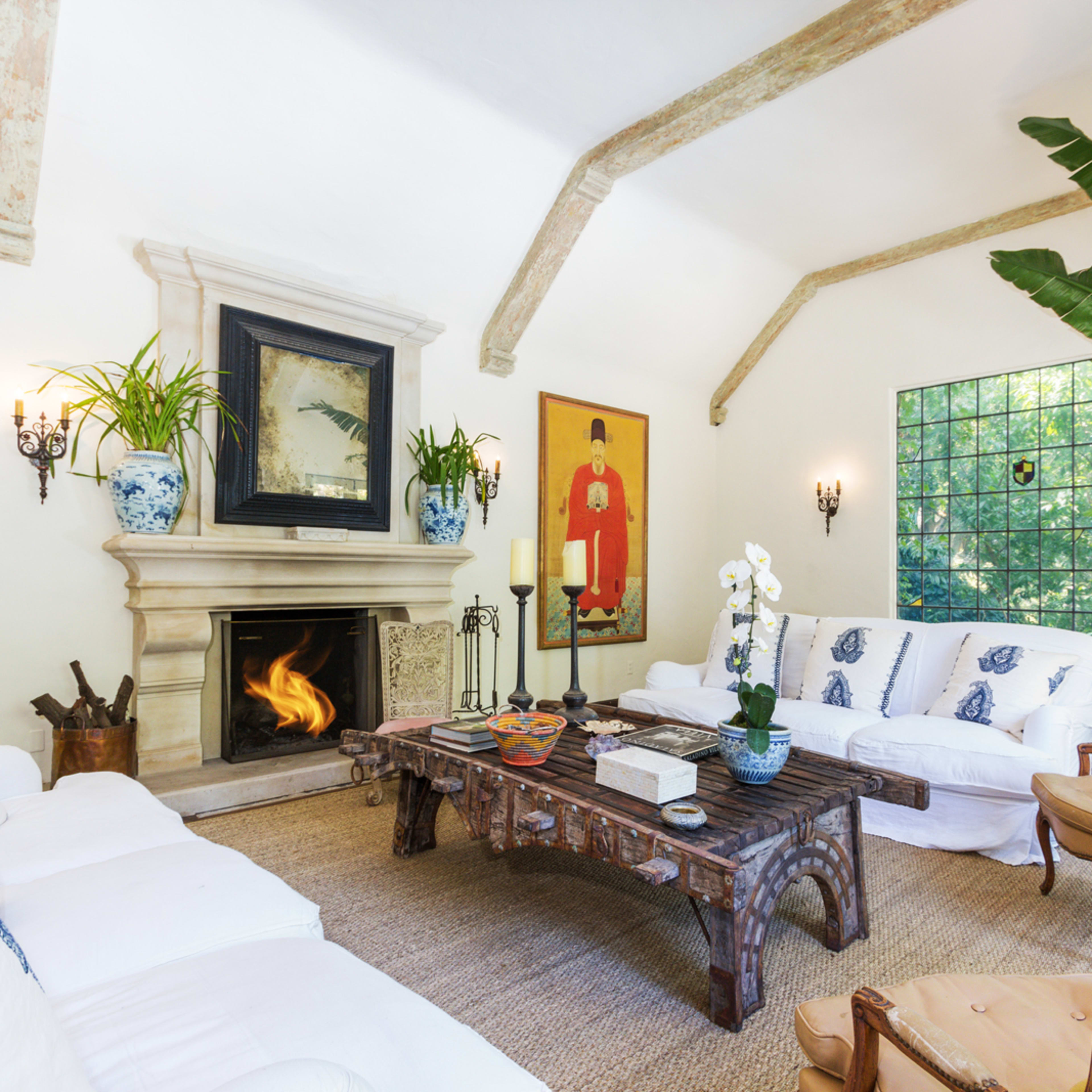 A spacious living room features a central coffee table in front of a white sofa, with a fireplace framed by artwork and potted plants decorating the space.