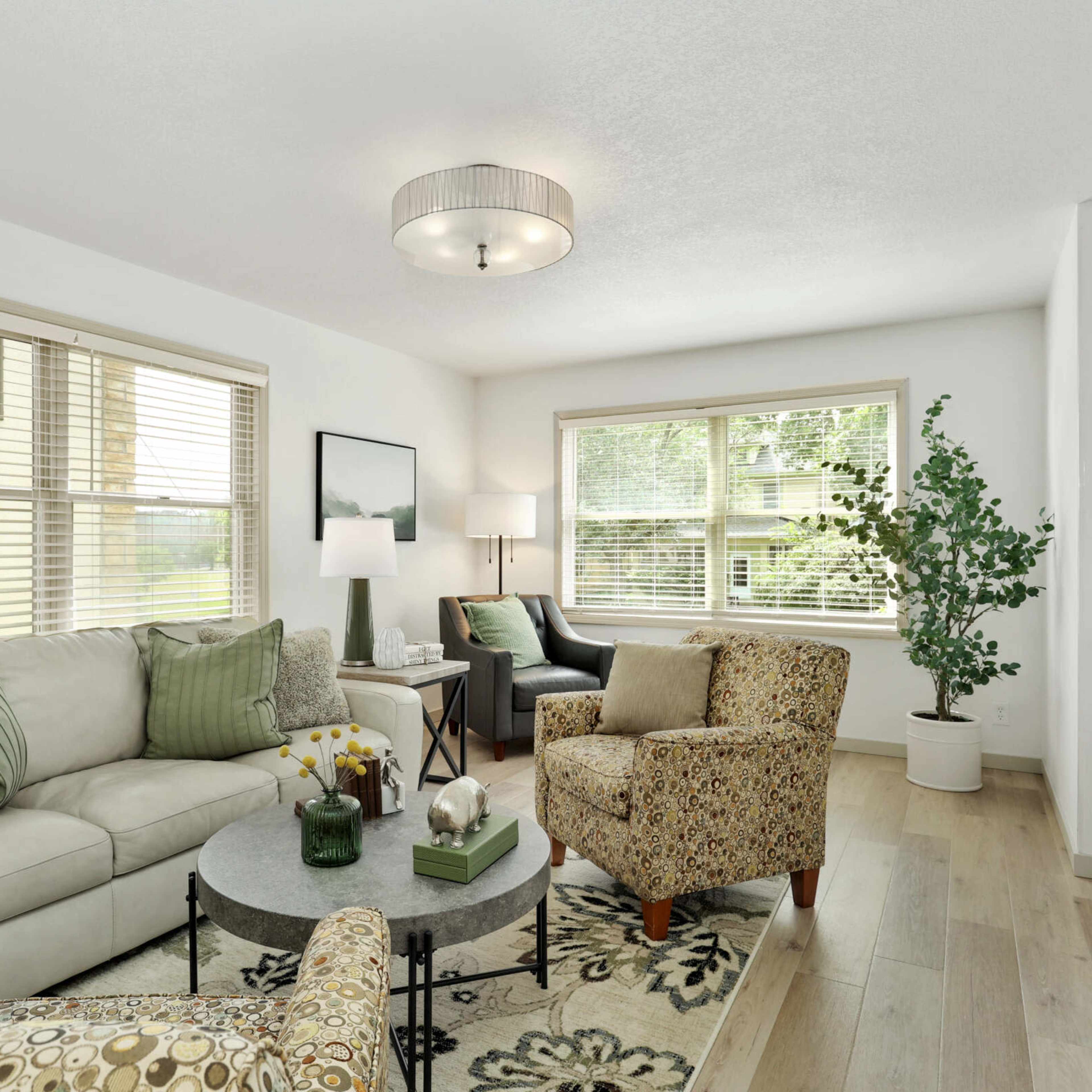 The living room features a light-colored sofa, a patterned armchair, a round coffee table, and large windows allowing natural light to fill the space.