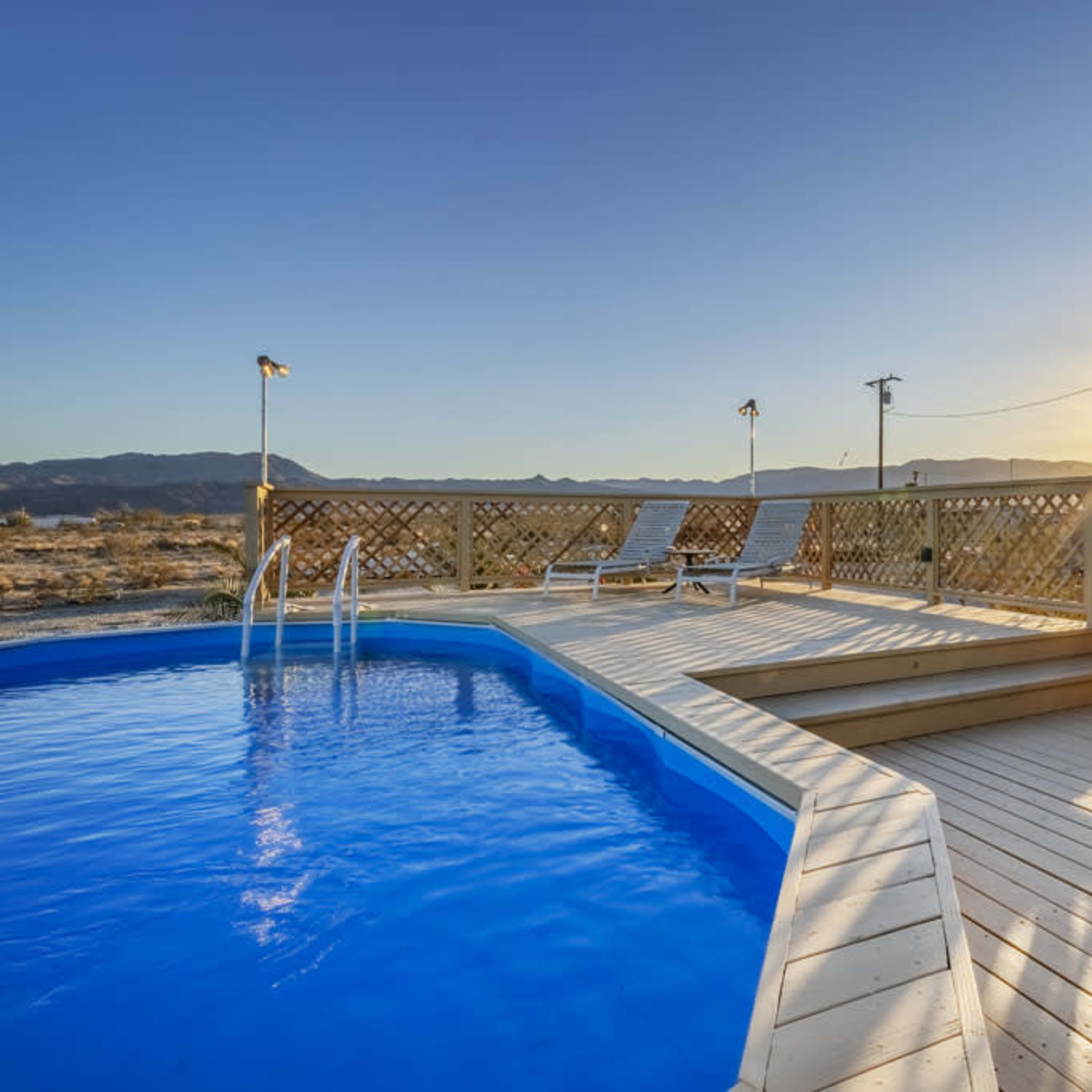 A clear blue pool is surrounded by a wooden deck, with a lounge chair positioned nearby, as the sun sets over distant mountains.