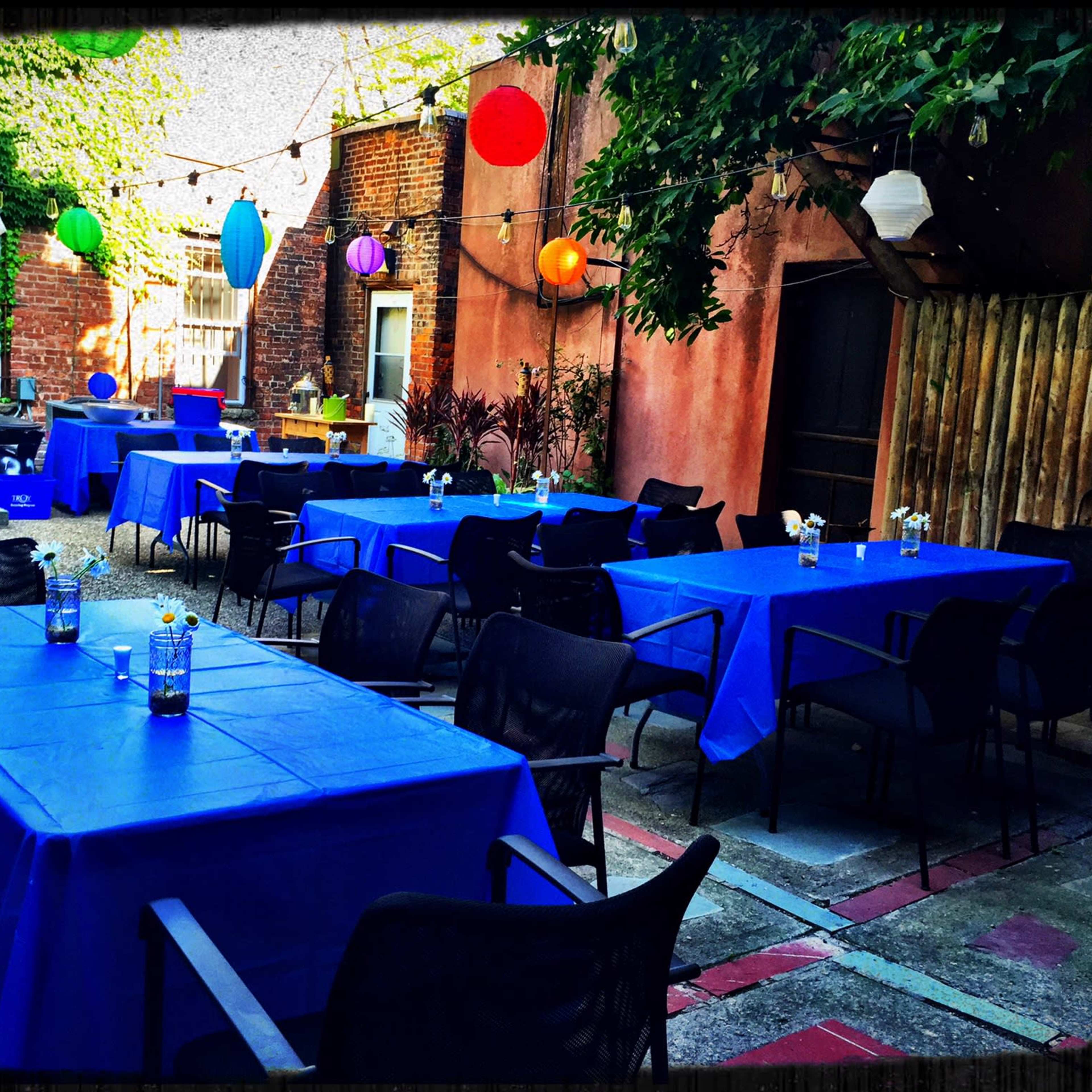 An outdoor area features several blue-covered tables arranged neatly, surrounded by colorful lanterns and string lights overhead.