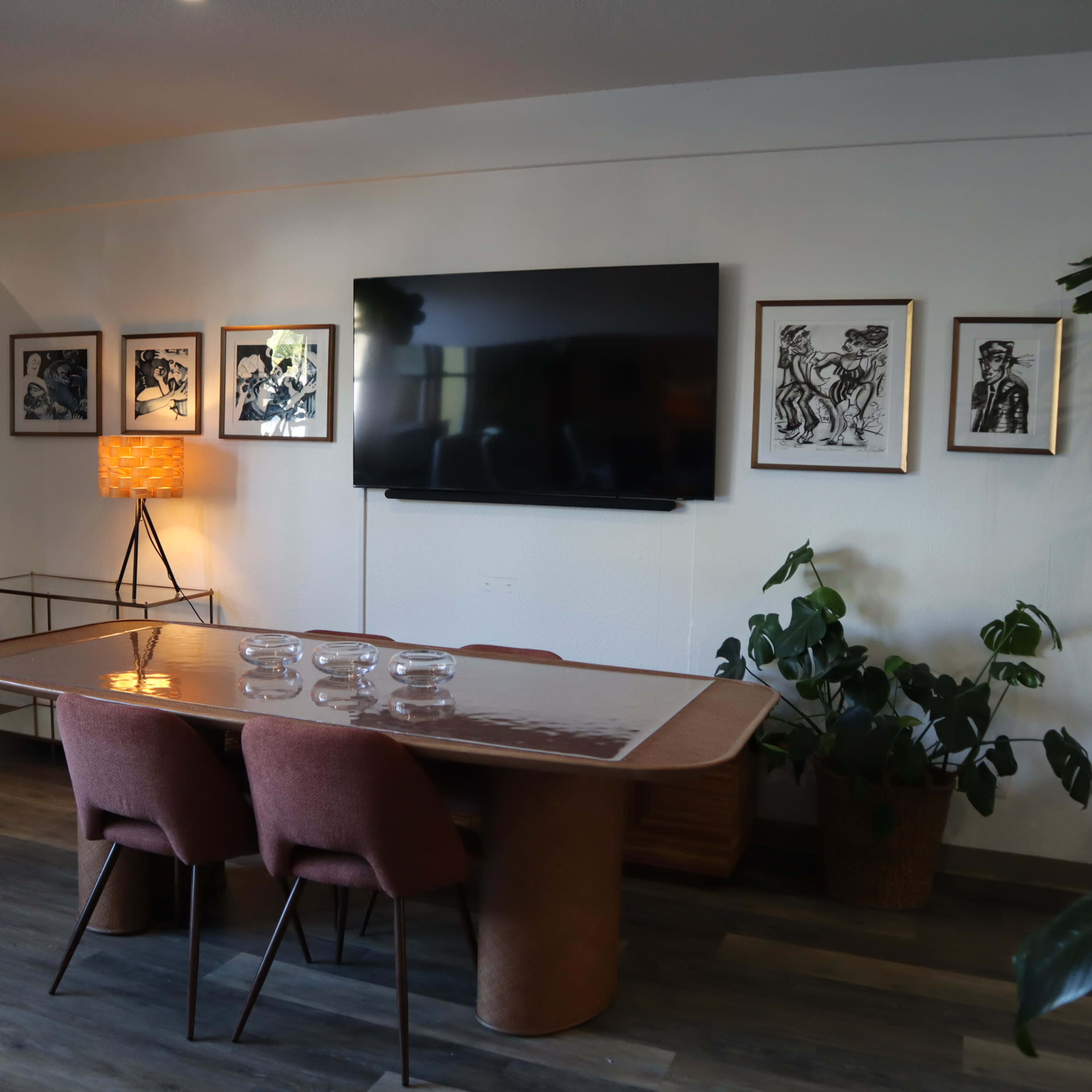 A modern conference room features a wooden table with pink chairs, surrounded by framed artwork on the walls and indoor plants.