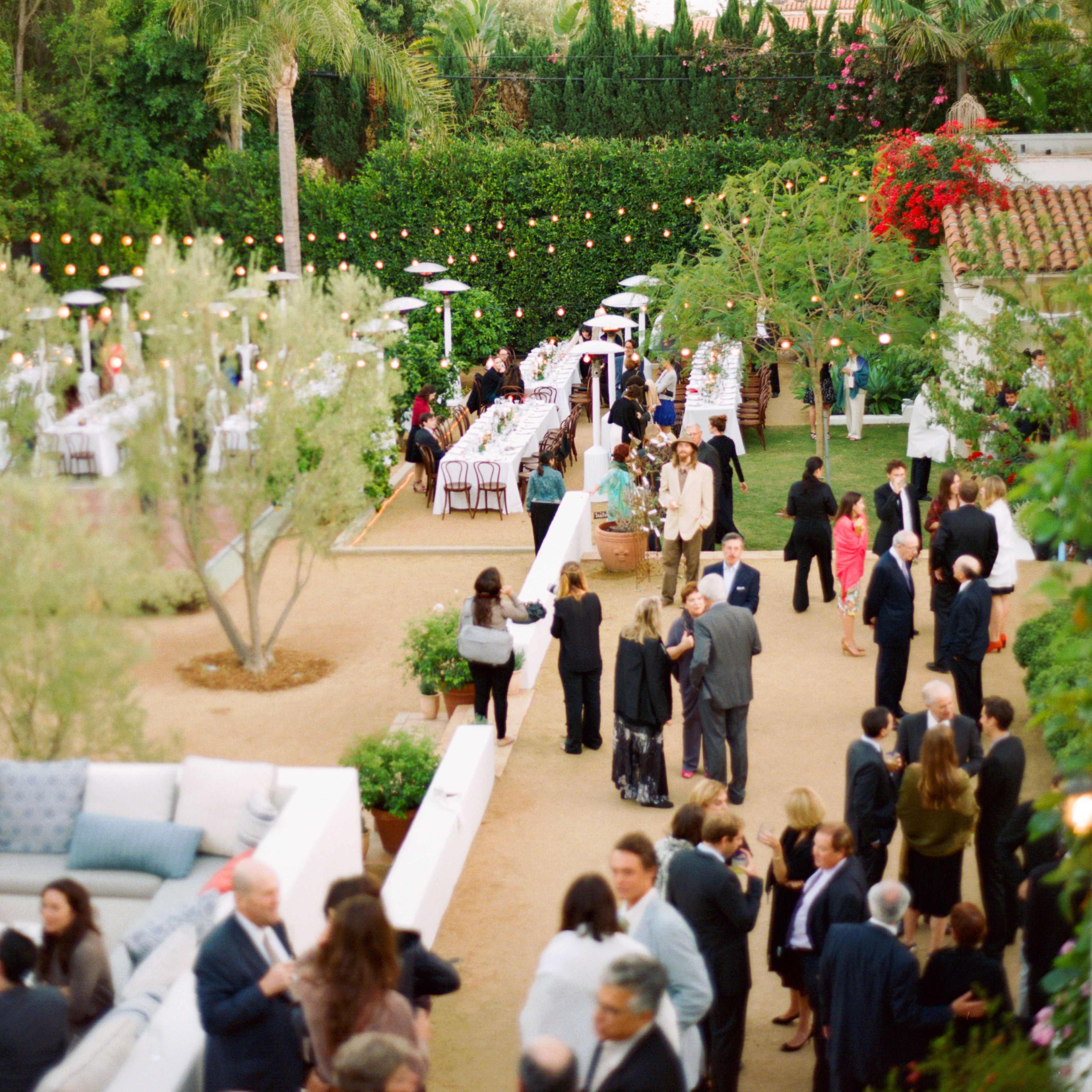 A garden party is set with tables and guests mingling amidst lush greenery and decorative lighting.