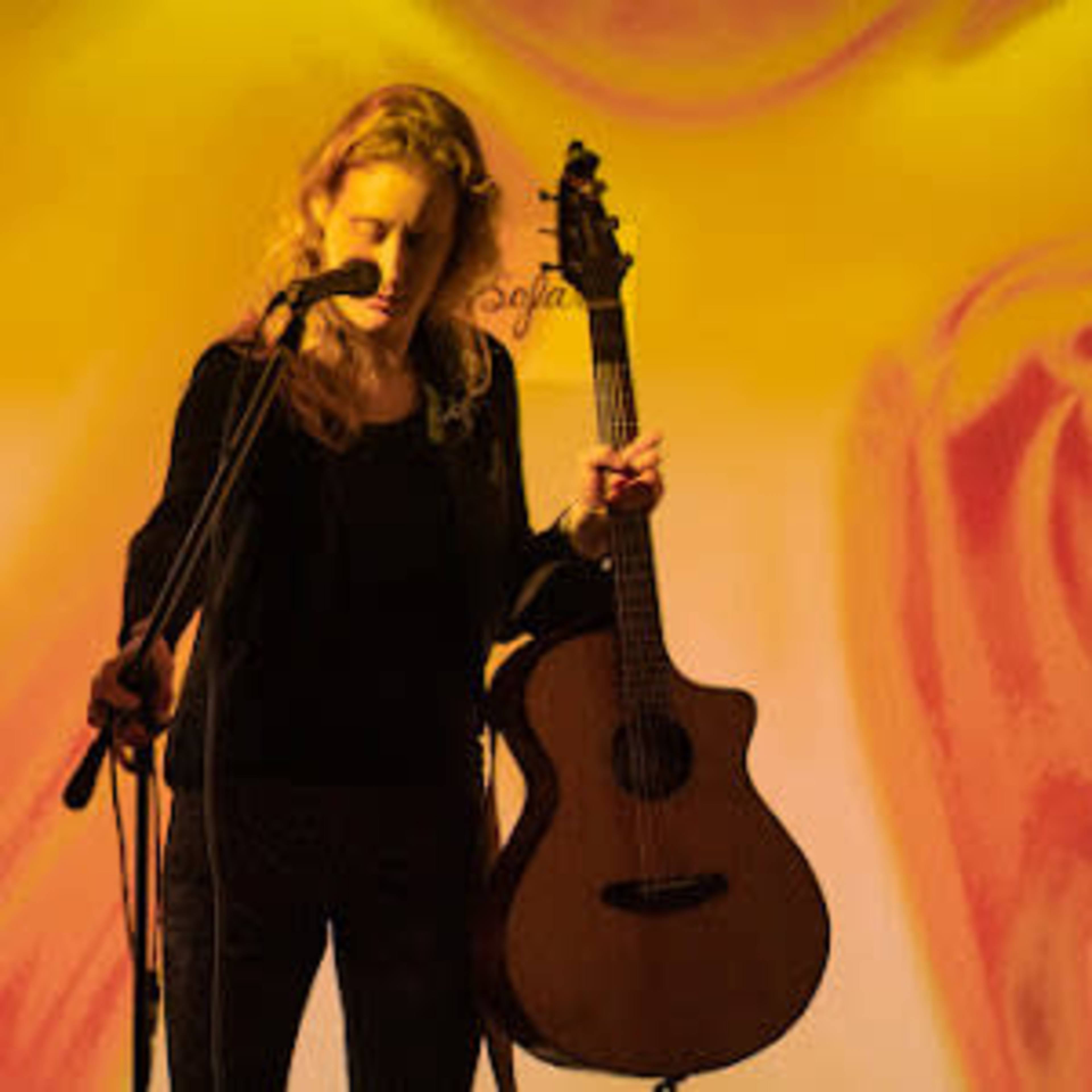 A woman stands with a guitar in hand, preparing to perform on a brightly colored stage.