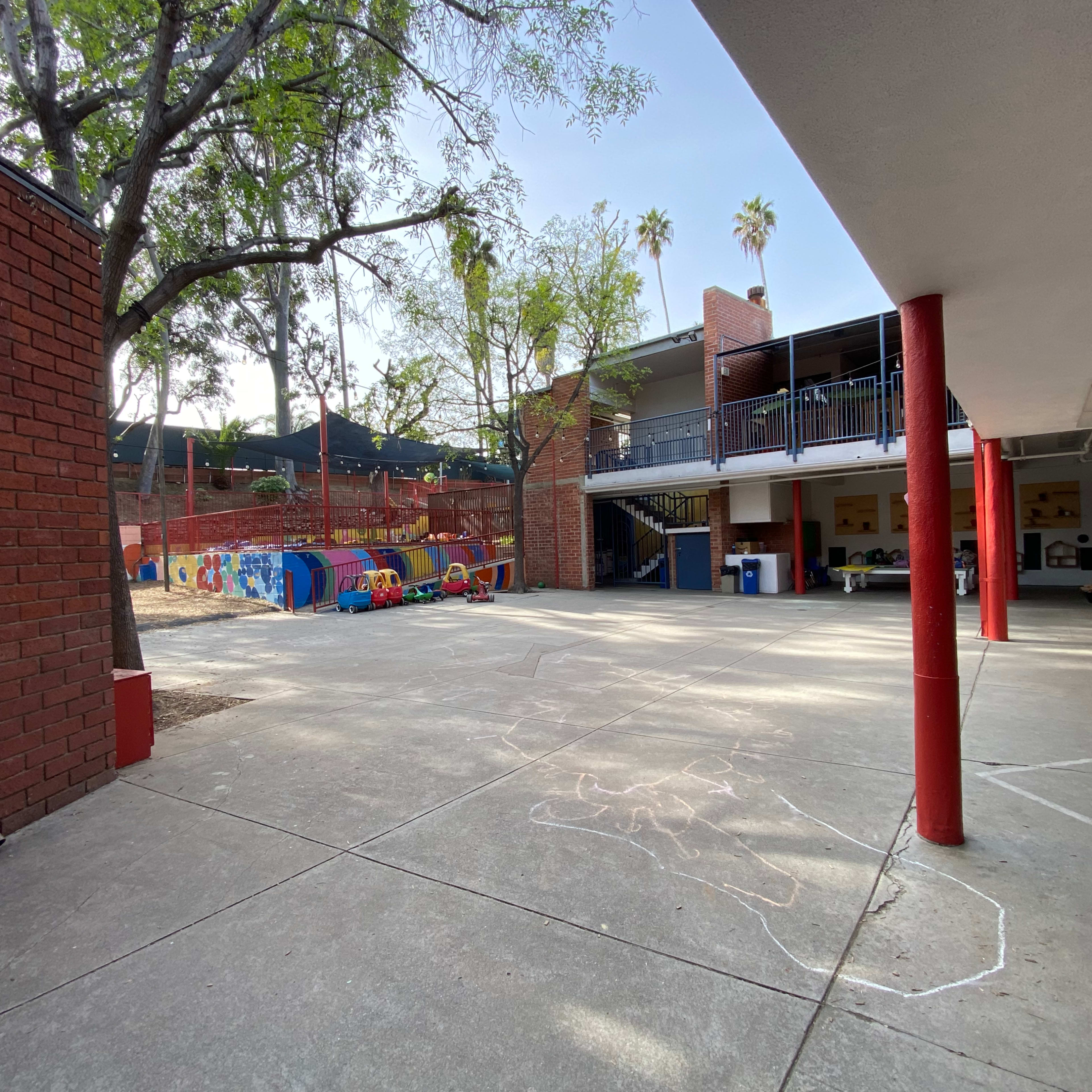A playground area with colorful equipment, surrounded by brick buildings and a concrete courtyard under a clear sky.